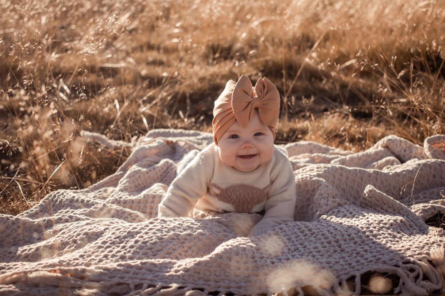 Baby in cream outfit smiles while lying on textured blanket in warm sunlight.