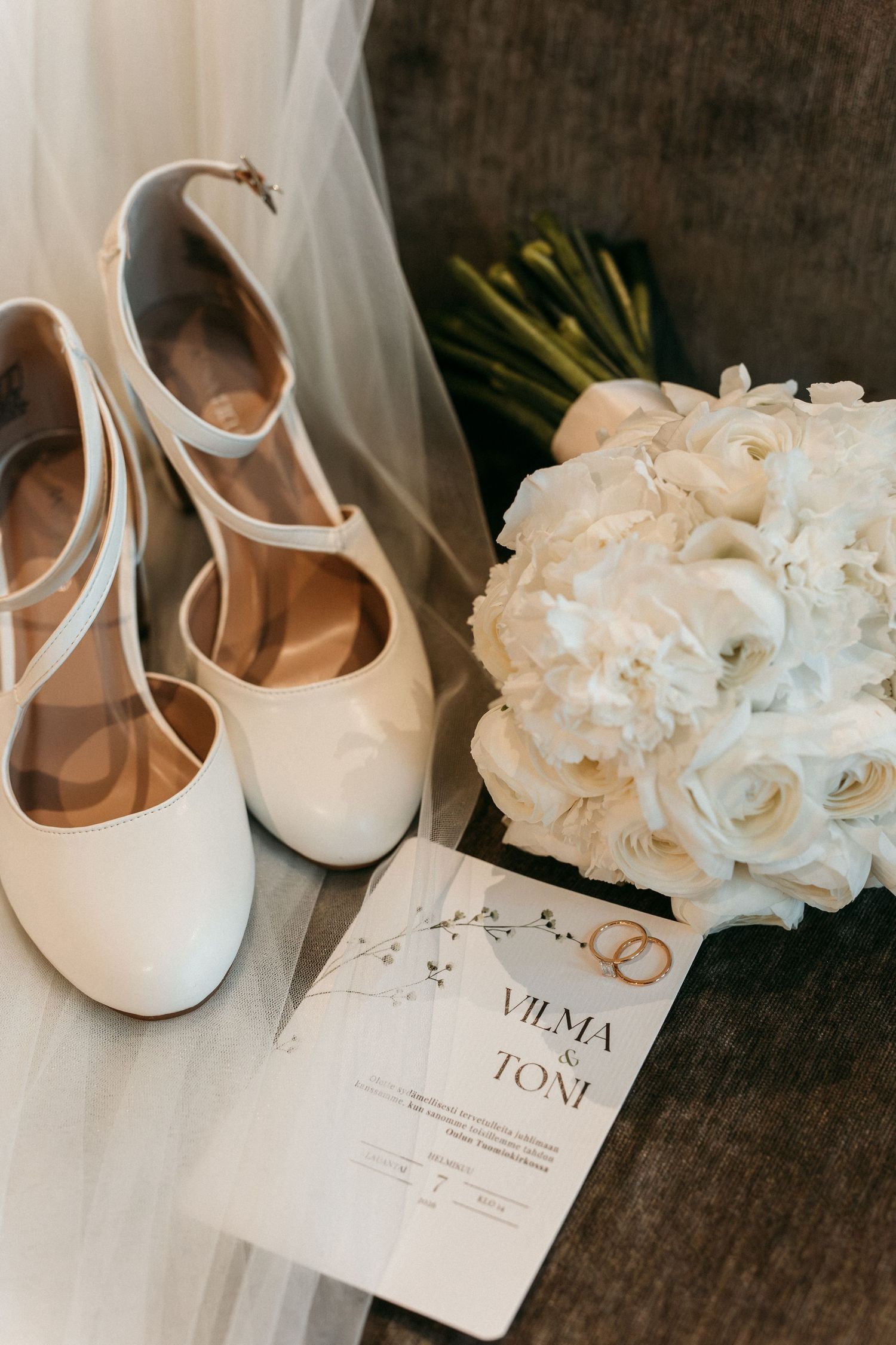 White bridal heels, wedding invitation for Vilma and Toni, and a white rose bouquet arranged elegantly on a gray surface.
