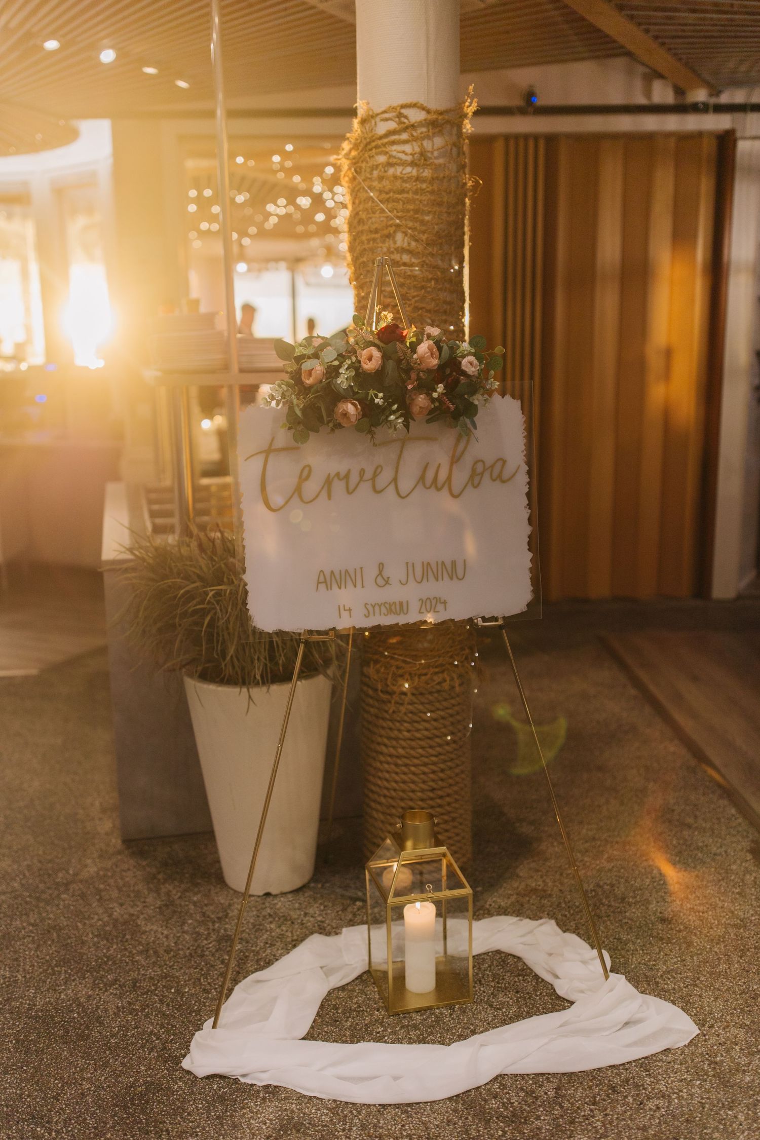 A rustic wedding welcome sign decorated with twinkle lights, florals, and fabric against a warm-lit backdrop.