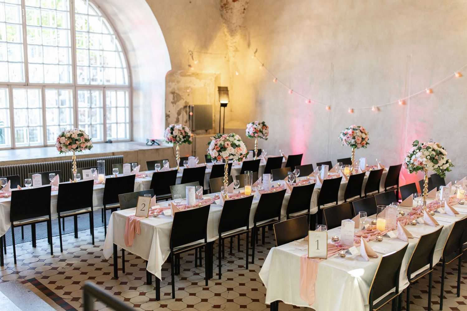 Modern wedding reception venue with long tables, black chairs, and pink accent lighting.
