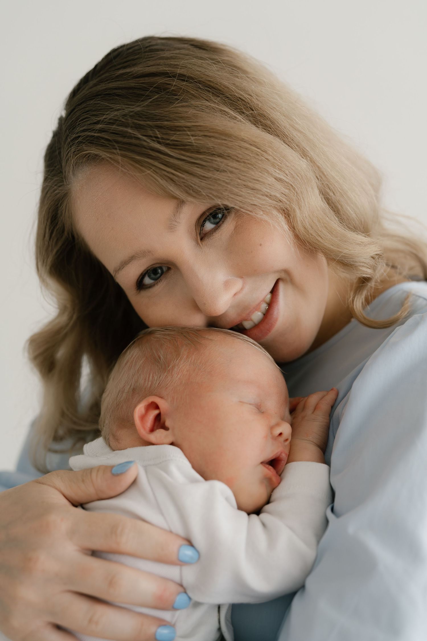 Smiling blonde mother tenderly holds her sleeping newborn baby close to her cheek against a white background.