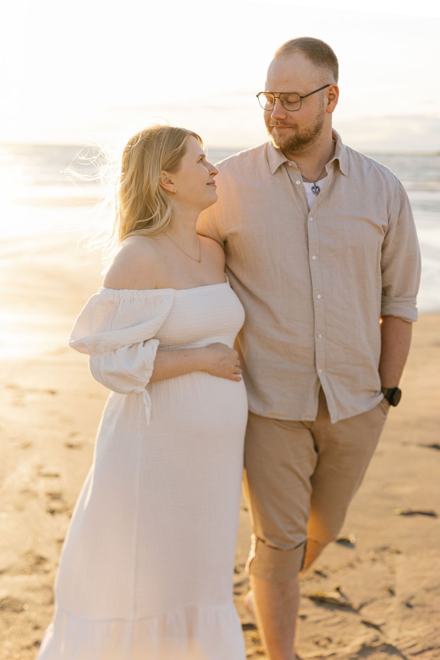 A romantic couple embraces on a sandy beach at sunset wearing white and beige clothing.