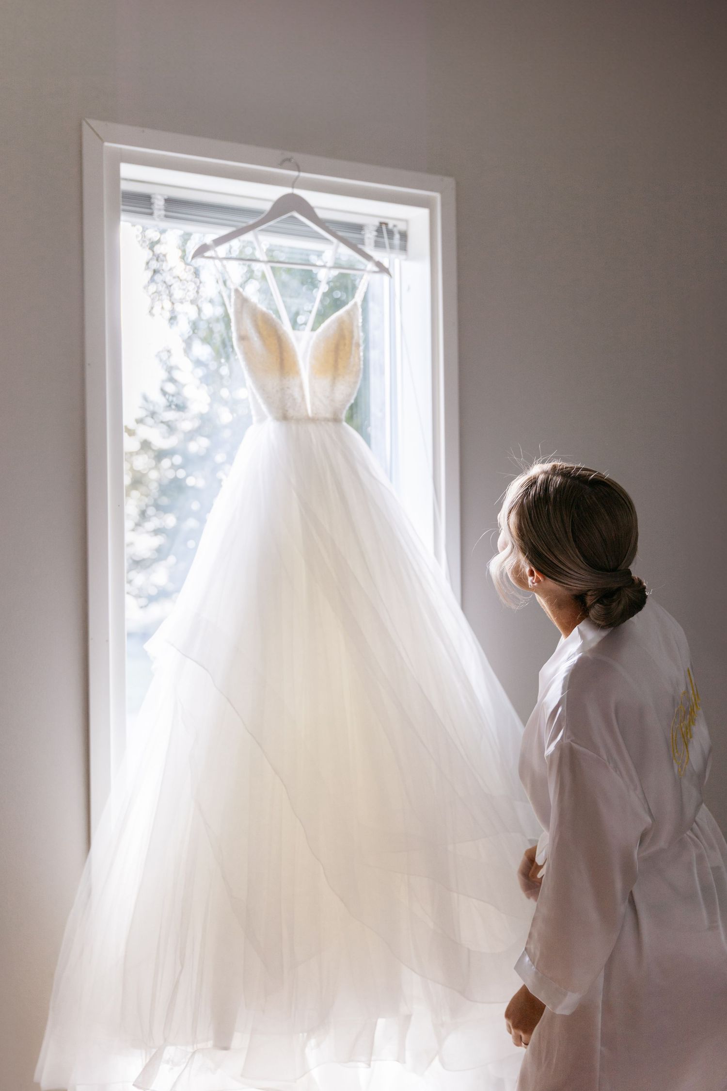 Wedding dress hanging in window light while figure in white robe admires it.
