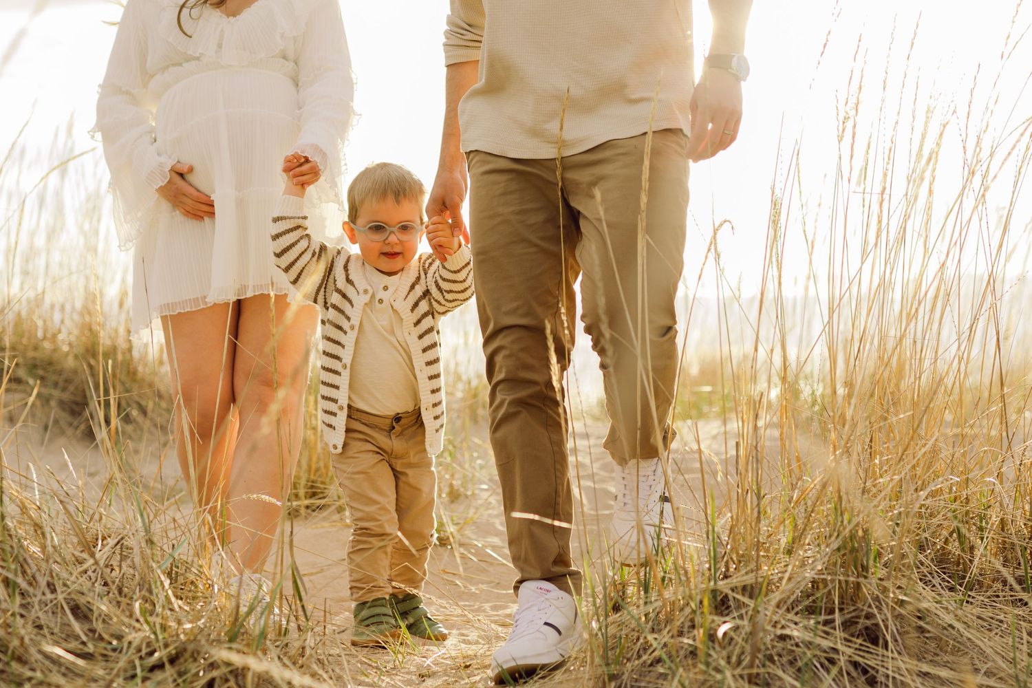 A family in coordinating neutral outfits walks together through tall golden grass at sunset.