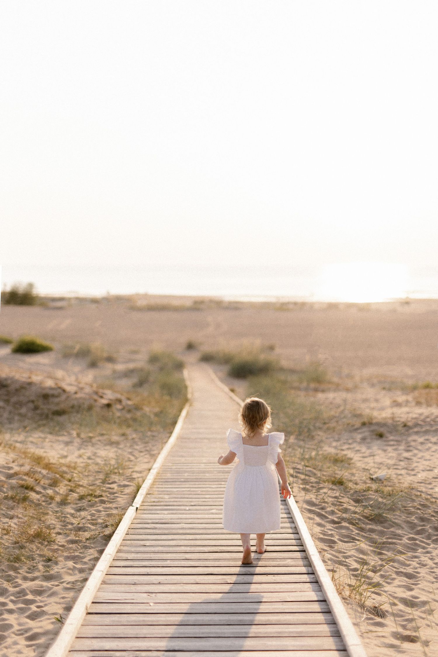 A small figure in white walks down a wooden boardwalk through sandy dunes at sunset.