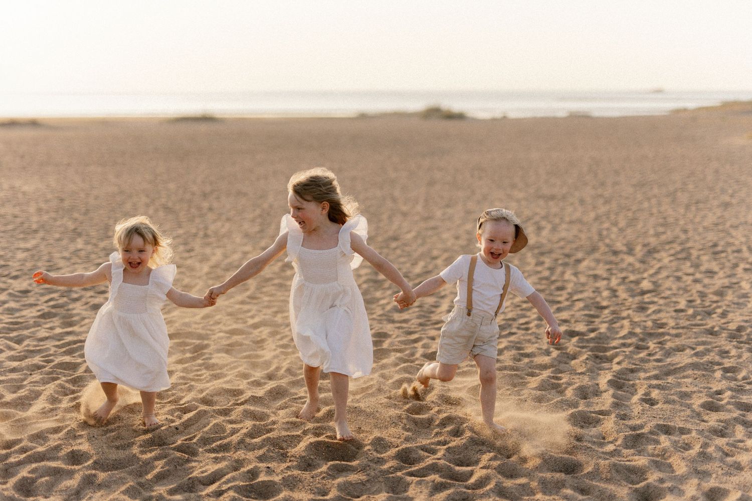 Three children holding hands run joyfully across a sandy beach at sunset, wearing matching white dresses.
