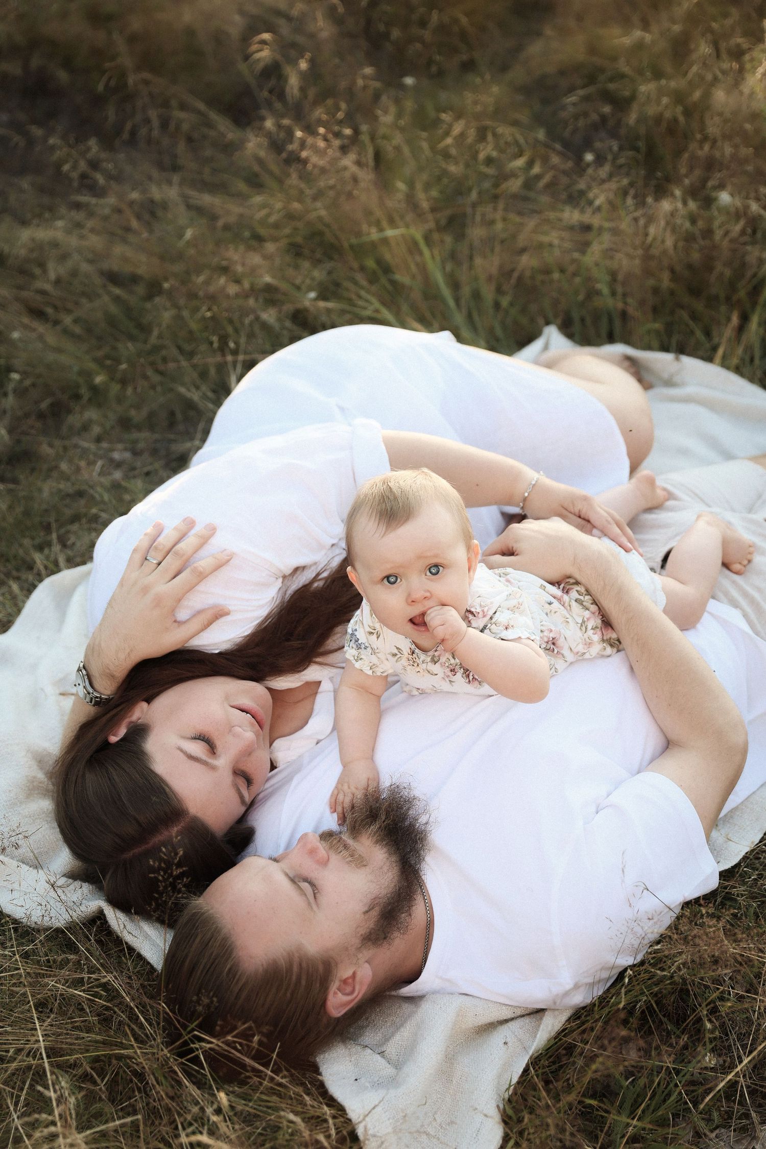 Family lying together on blanket in grassy field with baby resting on parent's chest.