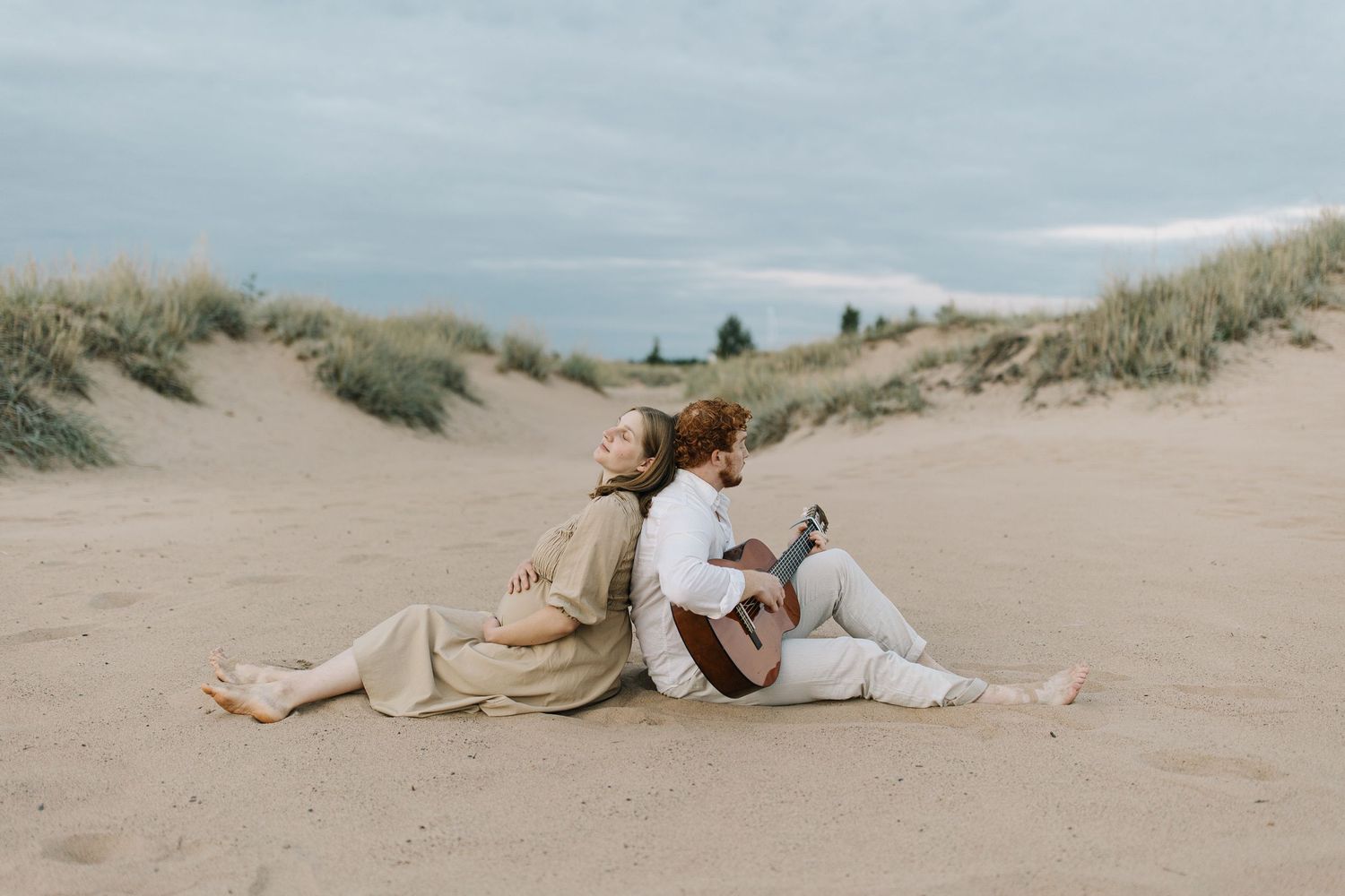 Couple sitting close together on blanket in sandy dunes near ocean.
