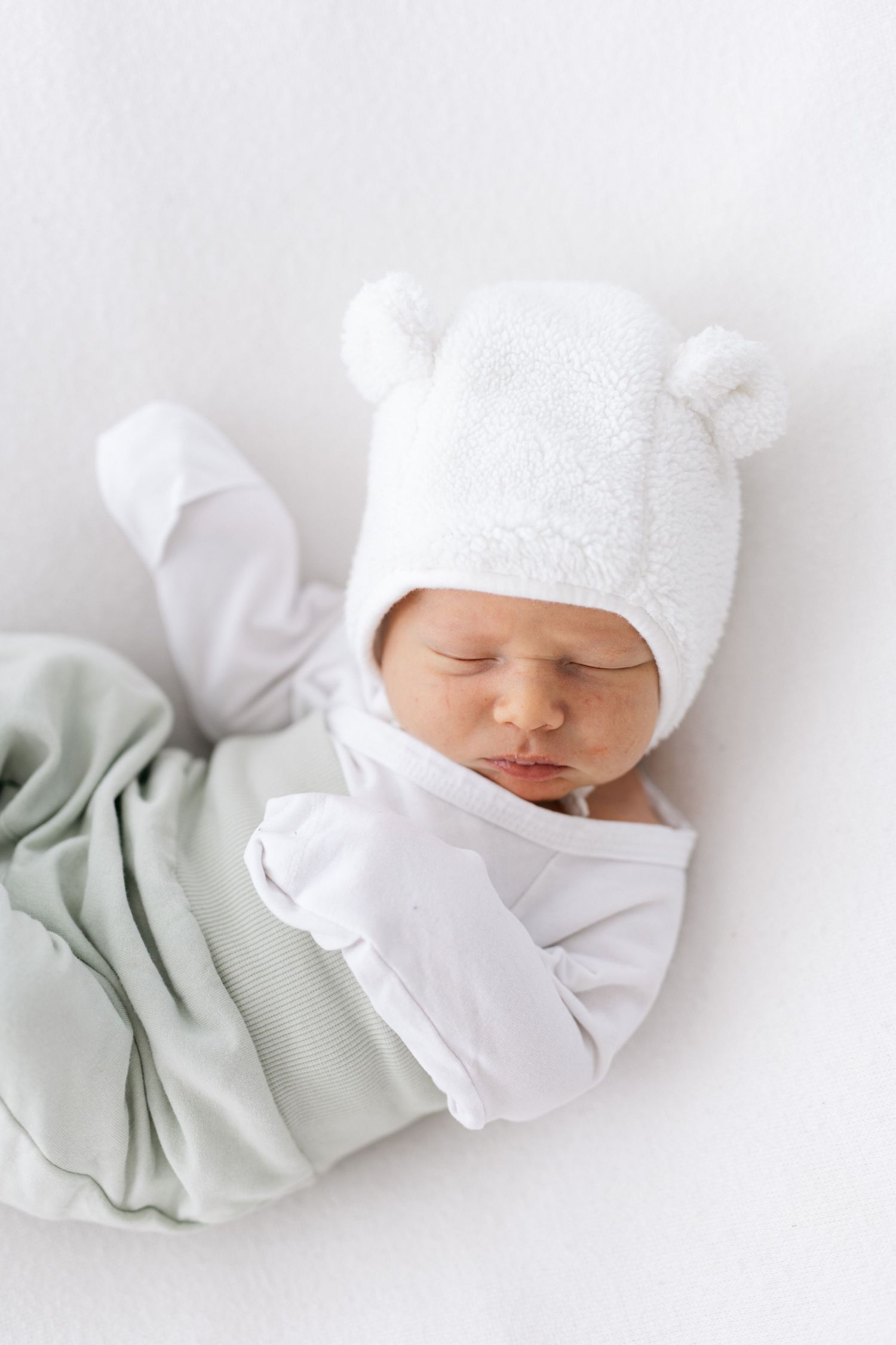 Sleeping newborn wearing white bear hat and swaddle on light background.