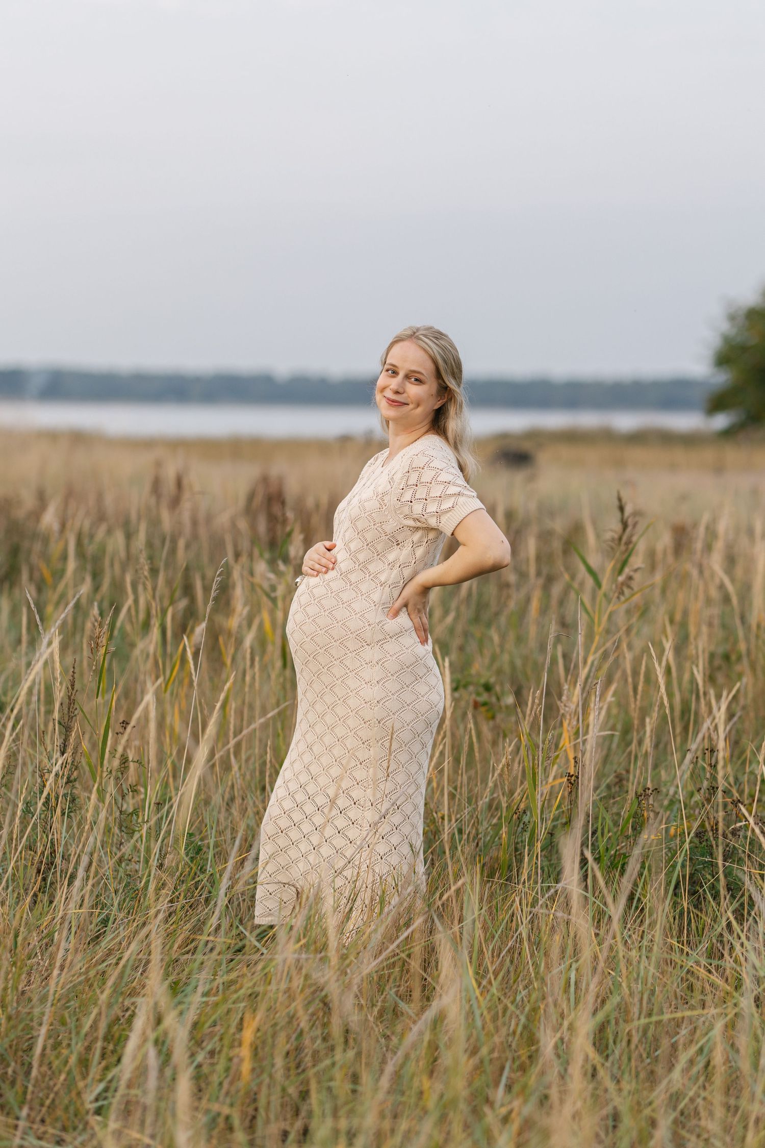 A pregnant woman in a lace dress stands in tall grass near a lake during golden hour.