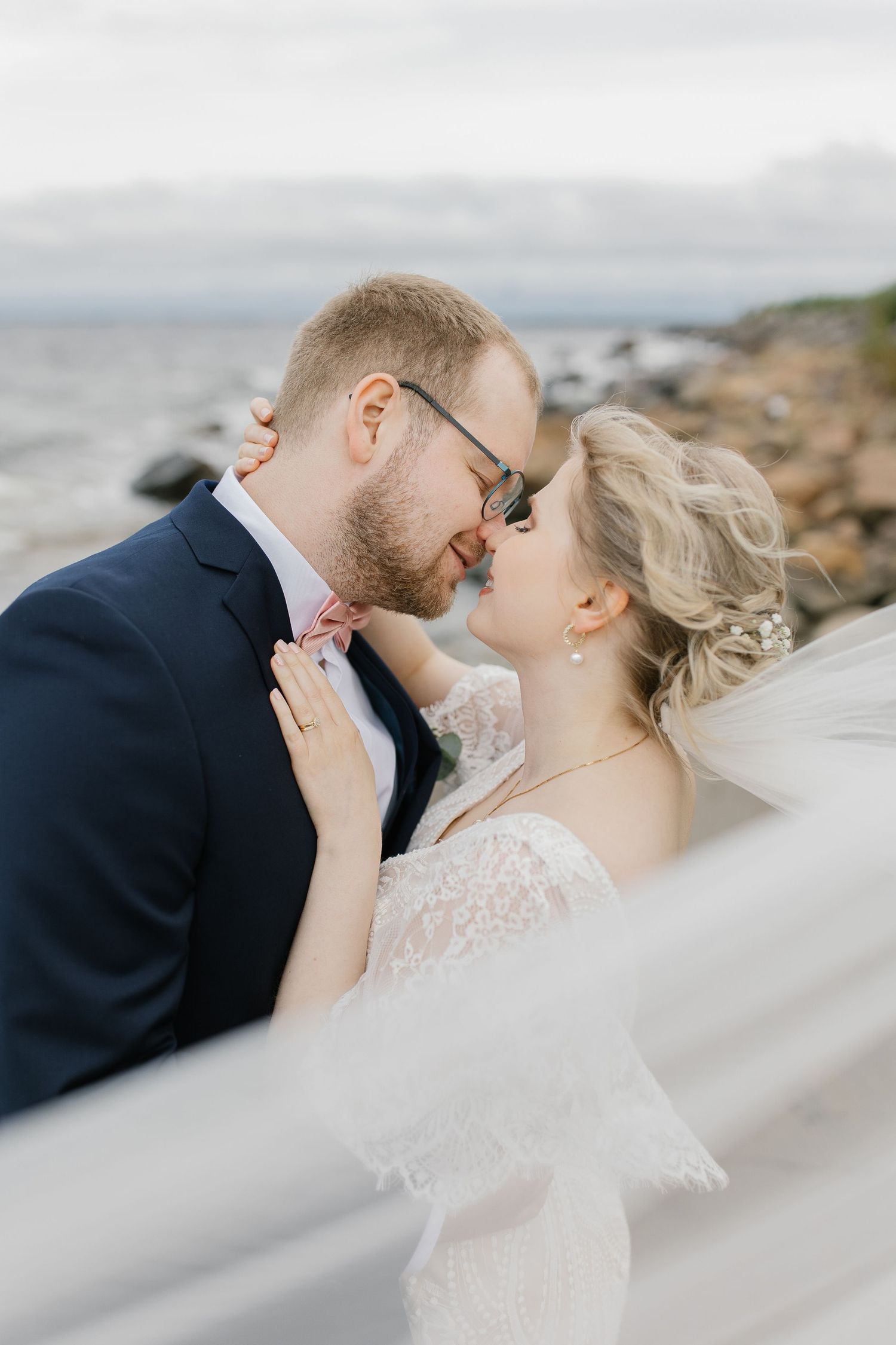 Romantic wedding portrait by rocky coastline with flowing veil in soft light.