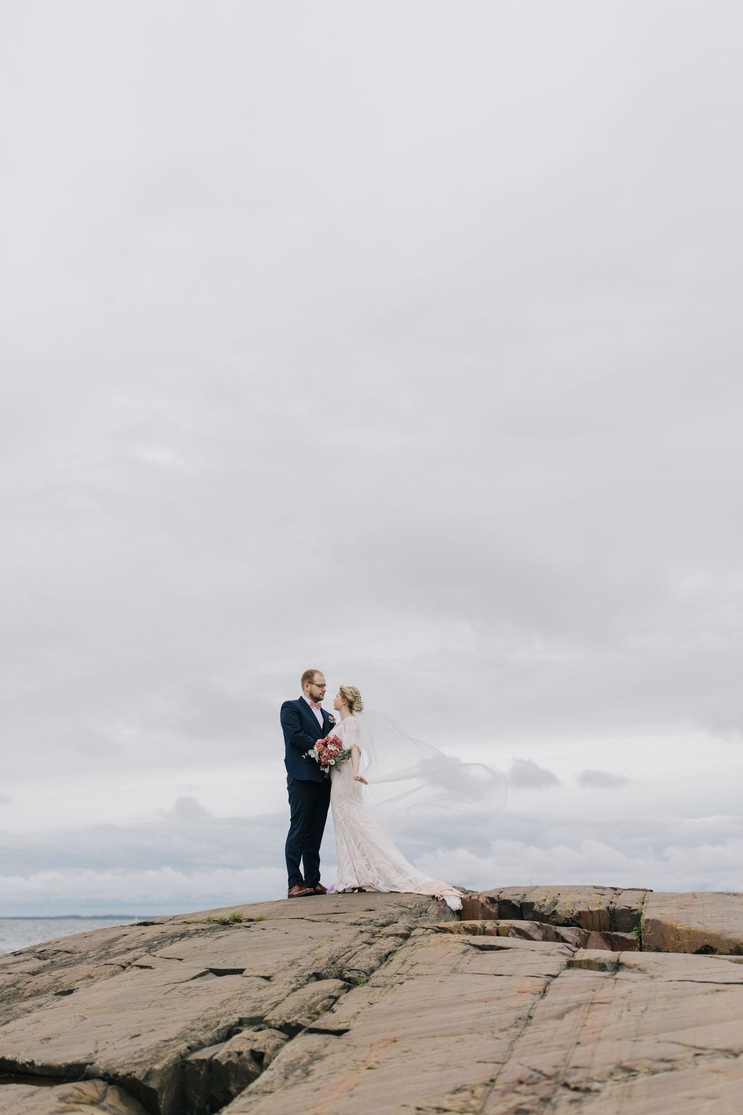 Wedding couple standing on dramatic rocky cliff overlooking ocean under cloudy sky.