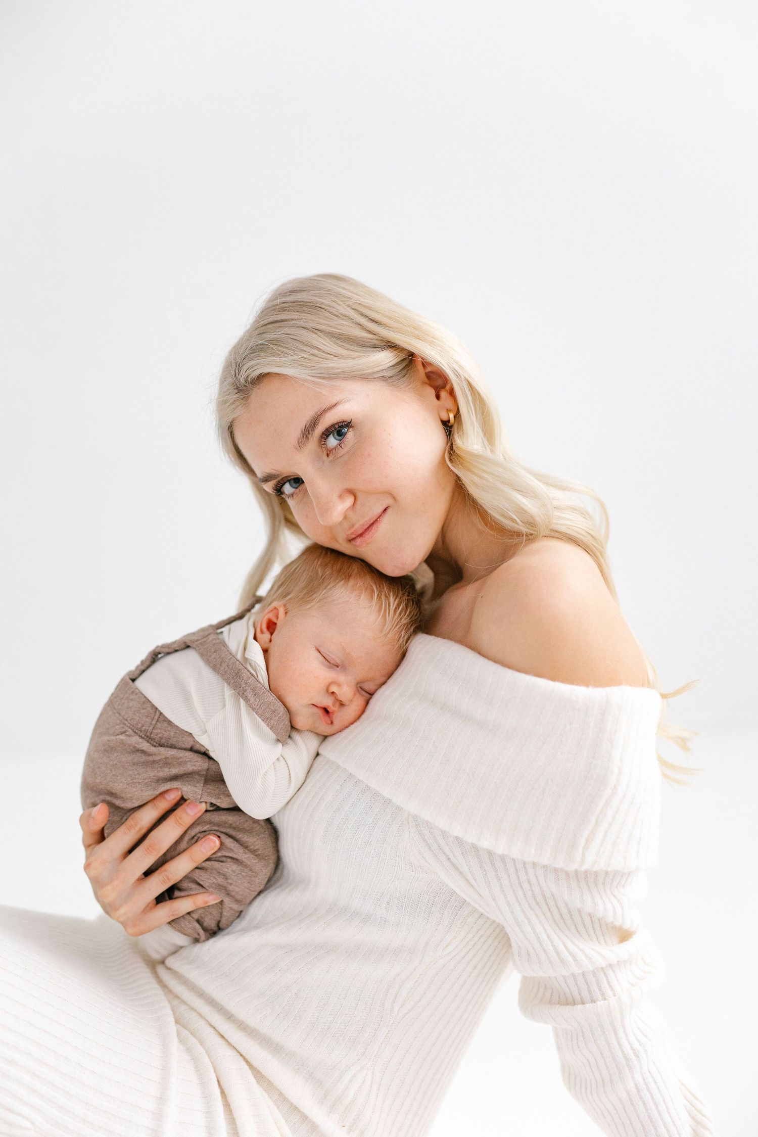 Parent wearing white sweater tenderly holds sleeping baby in brown outfit against white background.