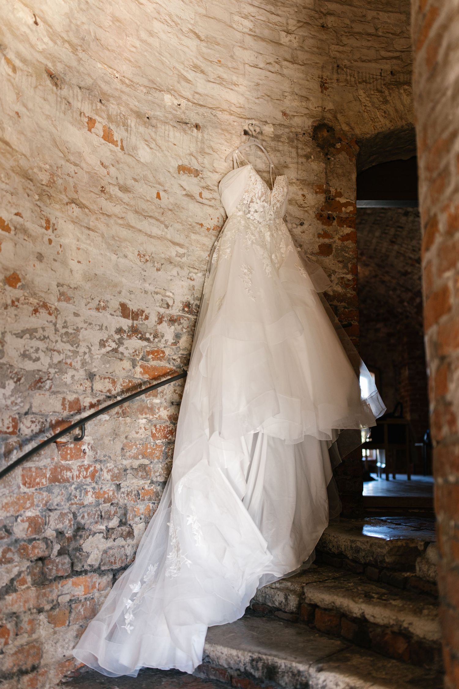 Elegant white lace wedding dress hanging on rustic brick wall in historic stairwell with dramatic lighting.