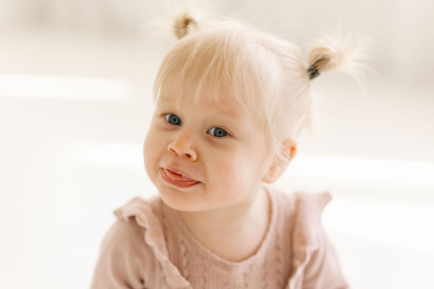 A small child with wispy blonde hair in pigtails smiles softly while wearing a light pink outfit.