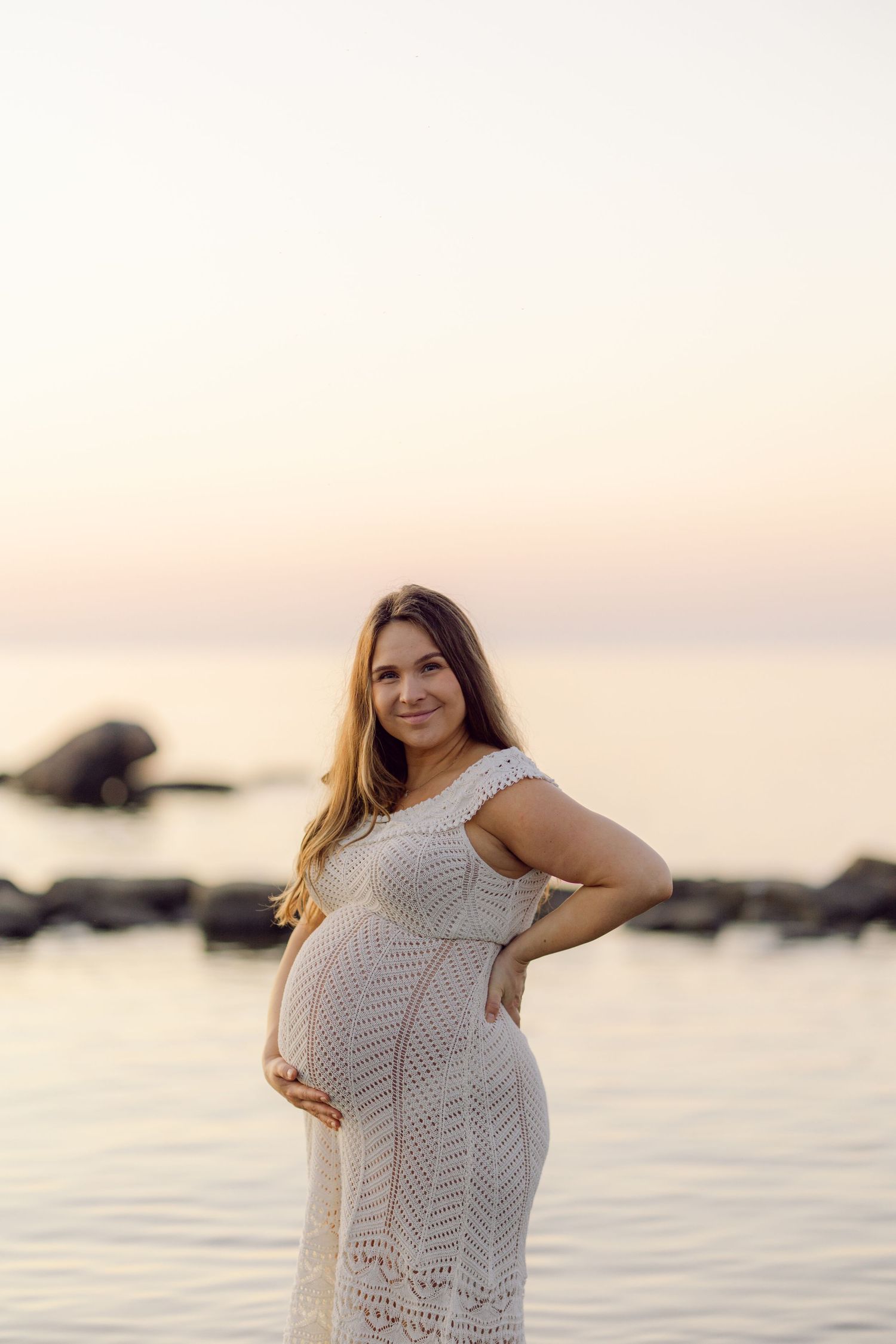 Expectant mother in white lace dress poses by the water during sunset.