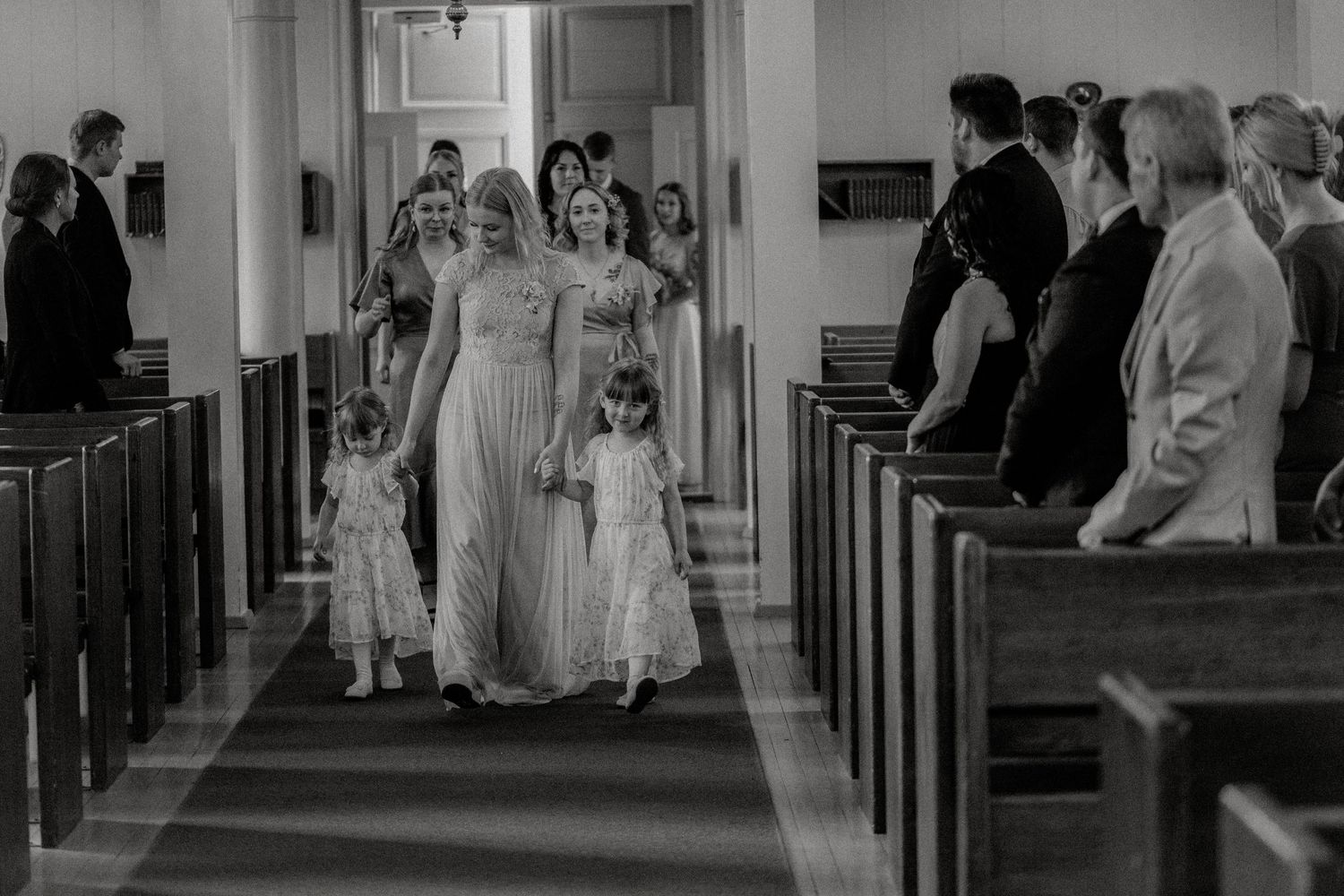 Wedding party walking down the church aisle in elegant white gowns while guests watch from the pews.