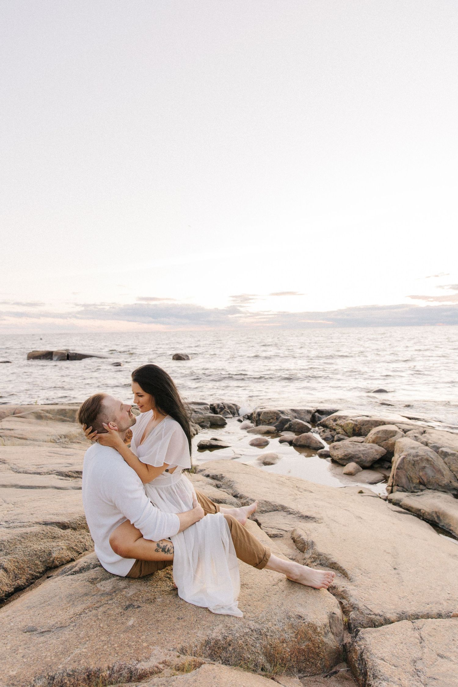 Romantic couple embracing on rocky beach at sunset in white outfits.
