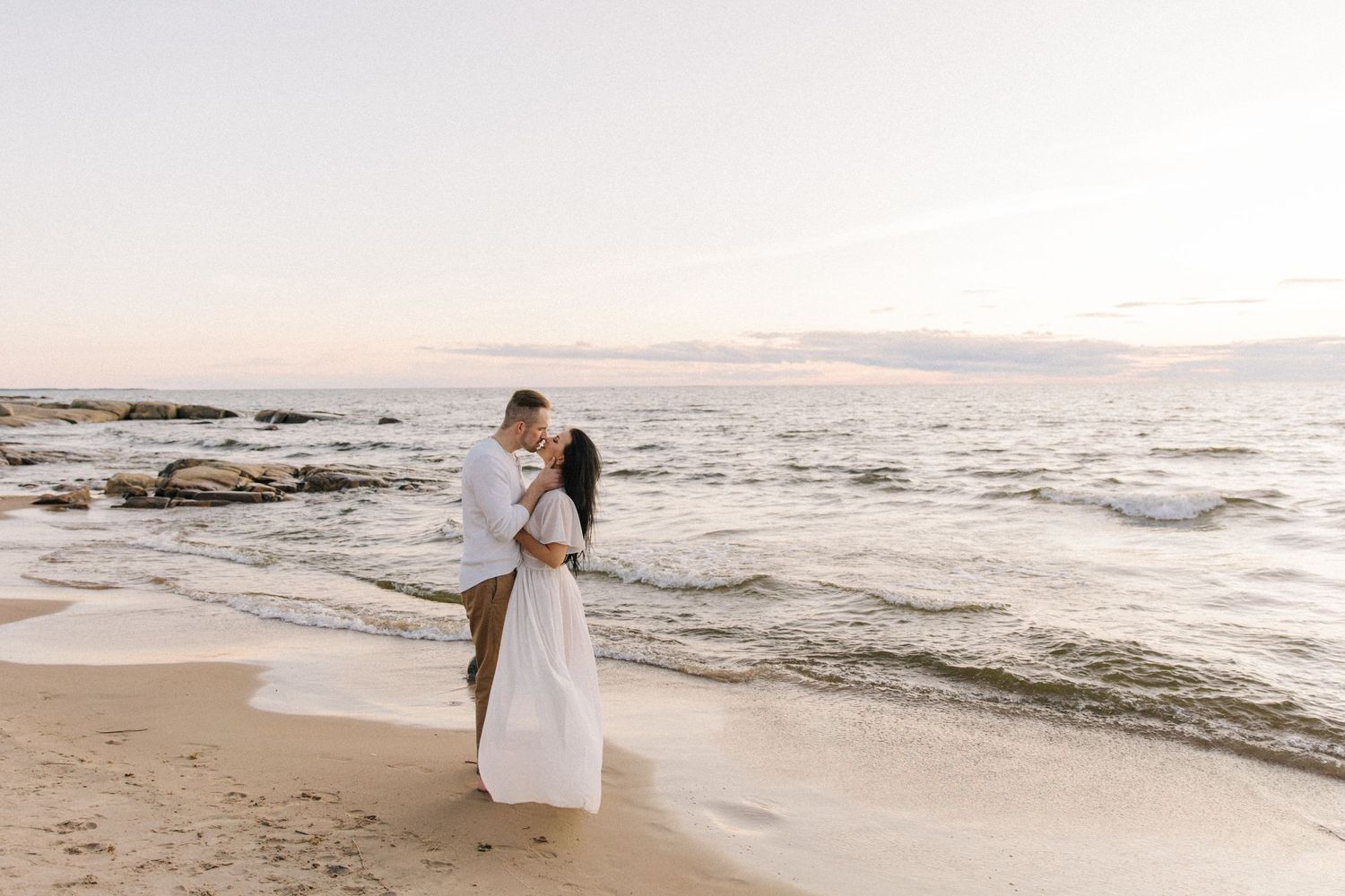 Romantic couple embraces on beach at sunset with waves crashing on rocky shore.