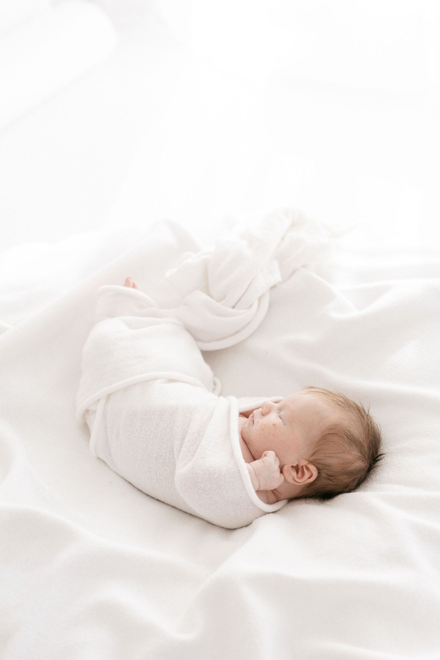 Newborn baby sleeping peacefully while swaddled in white on white bedding.
