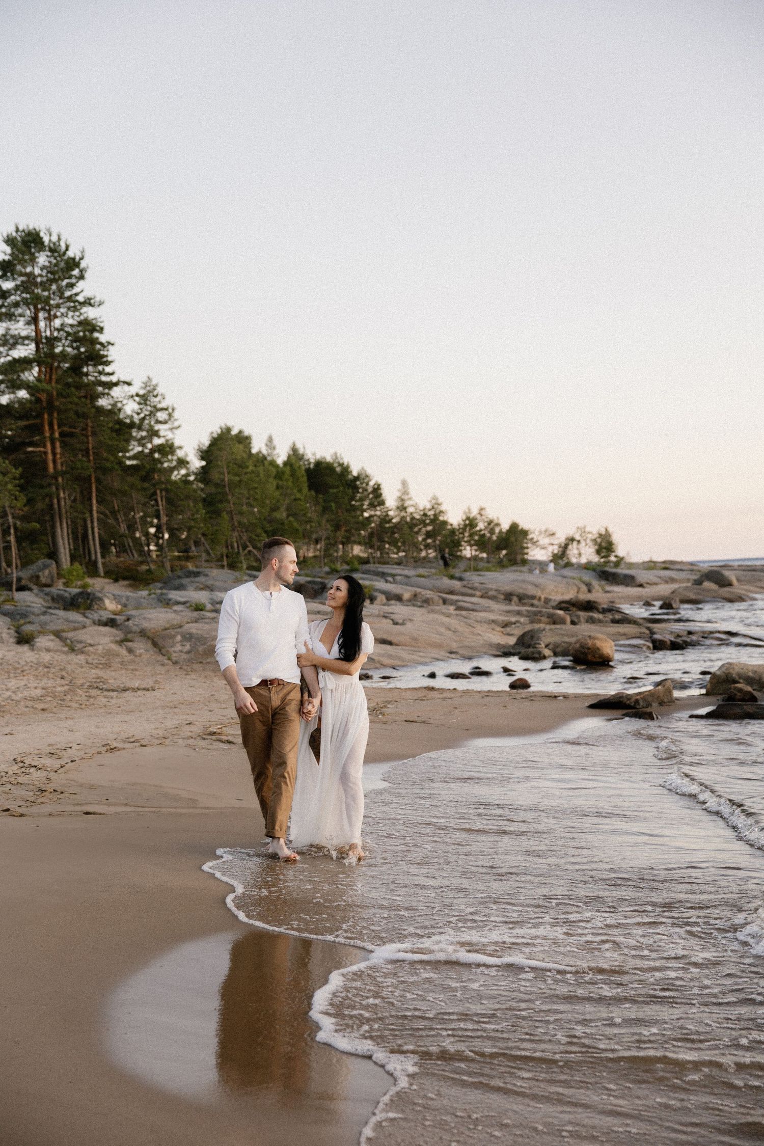 Couple walking hand in hand along a scenic beach with pine trees and rocky shore.