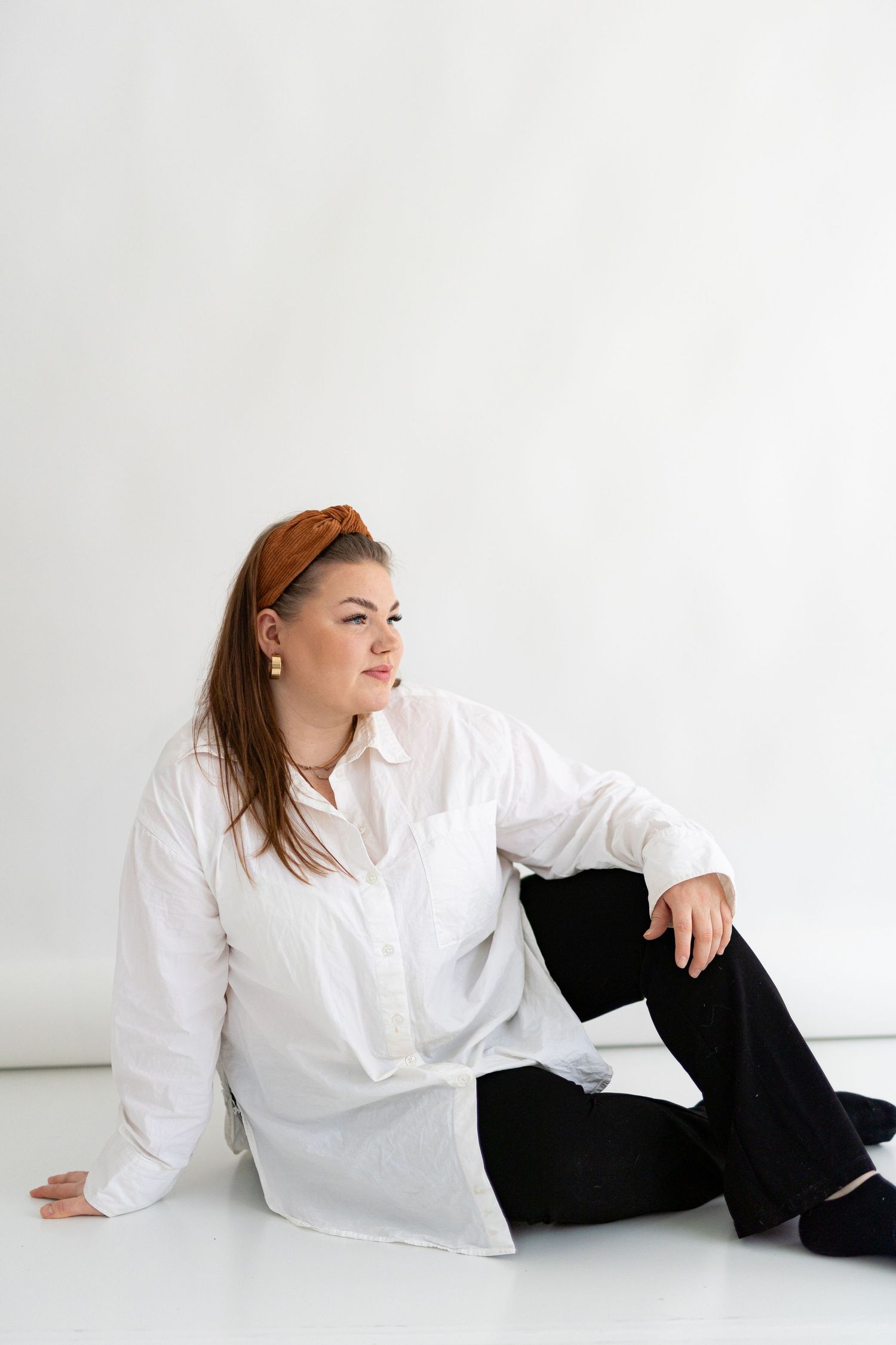 A person in white shirt and black pants sits casually on white floor against wall.
