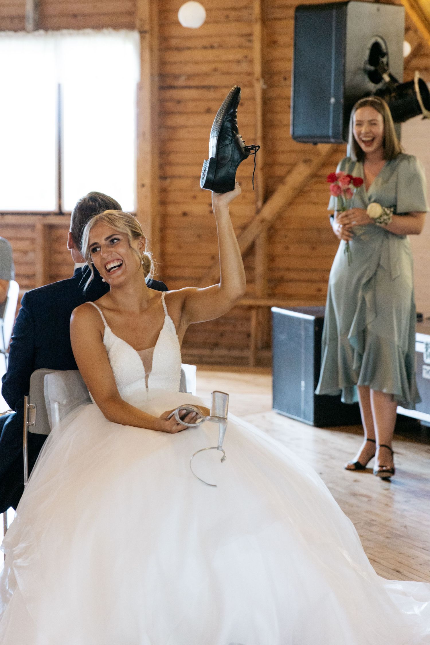 A joyful bride in a white gown playfully holds up a shoe while sitting at a rustic wooden venue.
