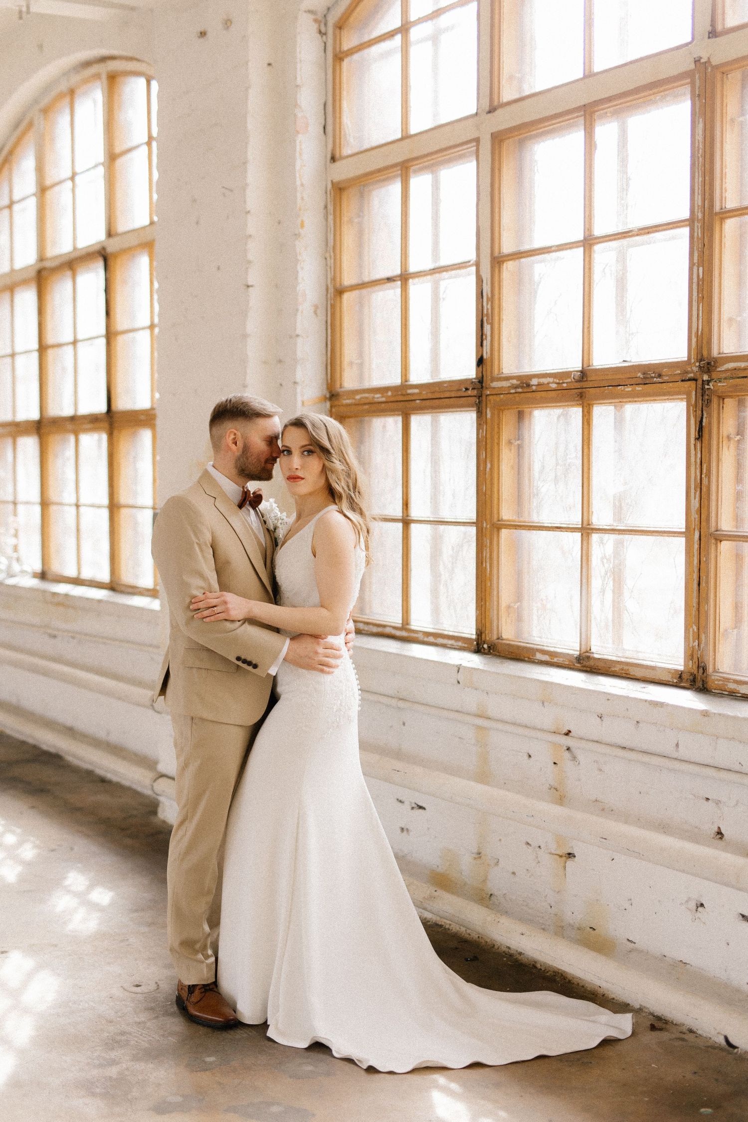 Elegant wedding couple standing together in white dress and tan suit by bright industrial windows.