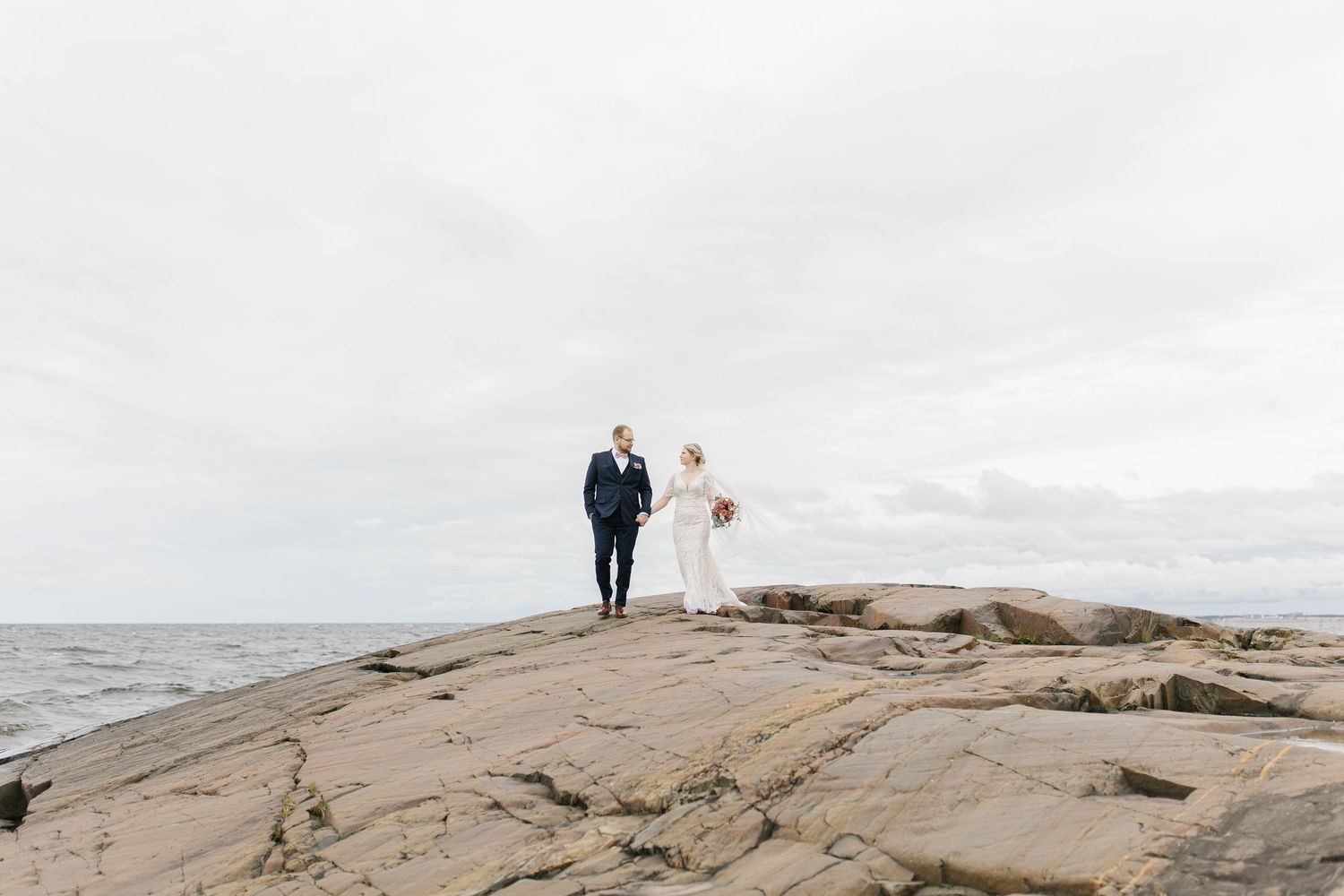Wedding couple stands atop rocky coastal outcrop overlooking ocean under overcast sky.
