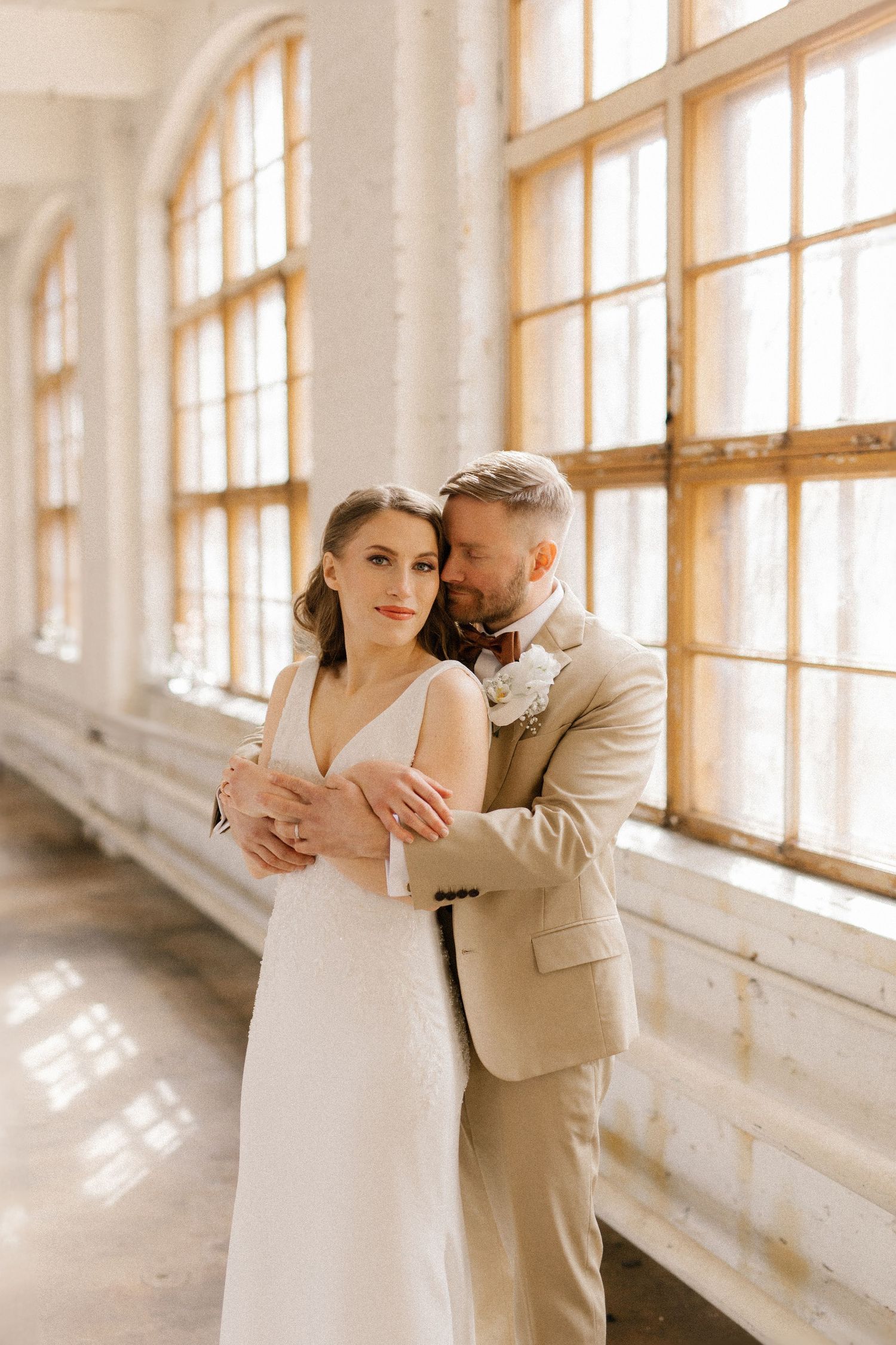Wedding couple share intimate moment in sunlit room with large windows.