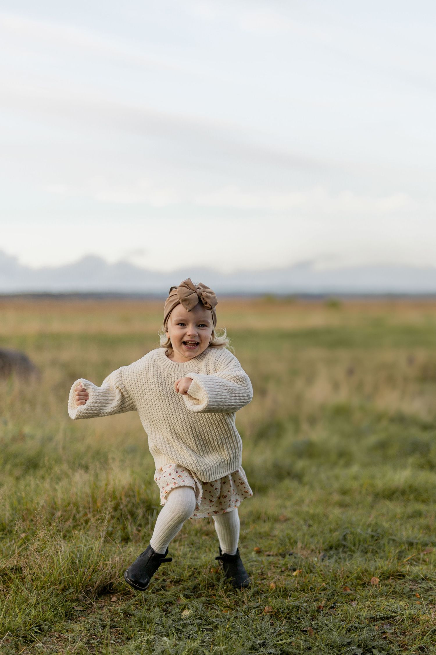 Small child wearing a cream sweater runs joyfully through a grassy field.