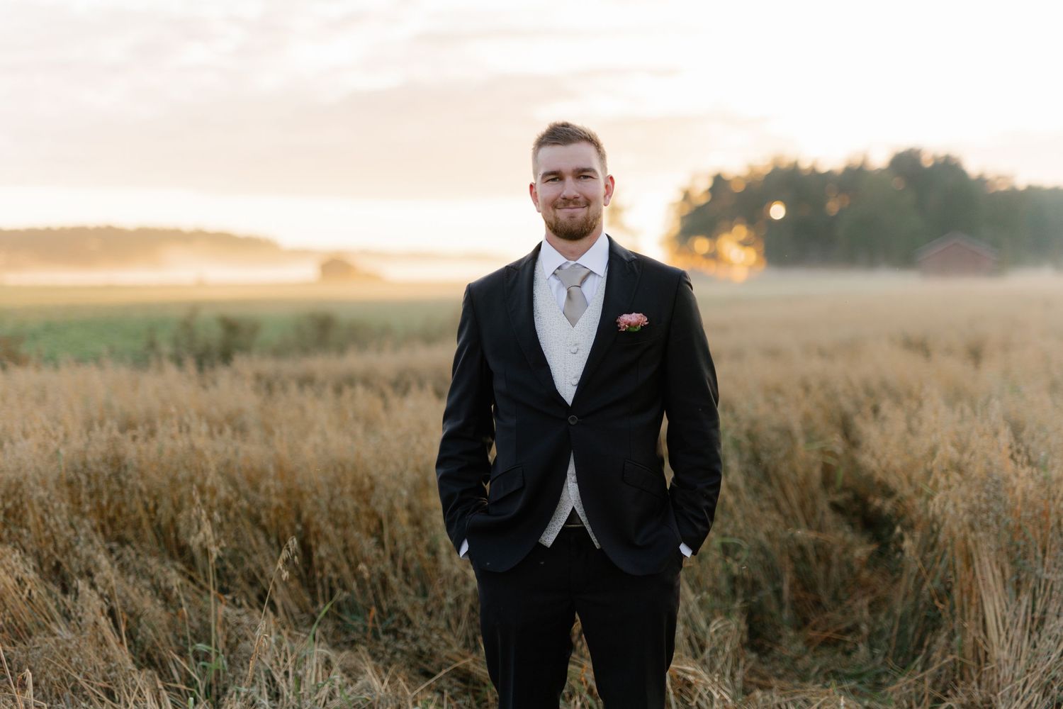 Groom in black suit standing alone in golden field at sunset.