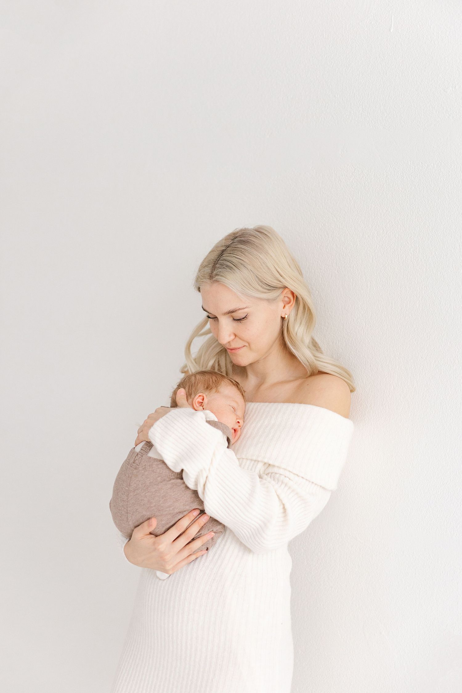 A tender moment captured as a parent in a white off-shoulder dress holds and gazes down at their sleeping newborn.