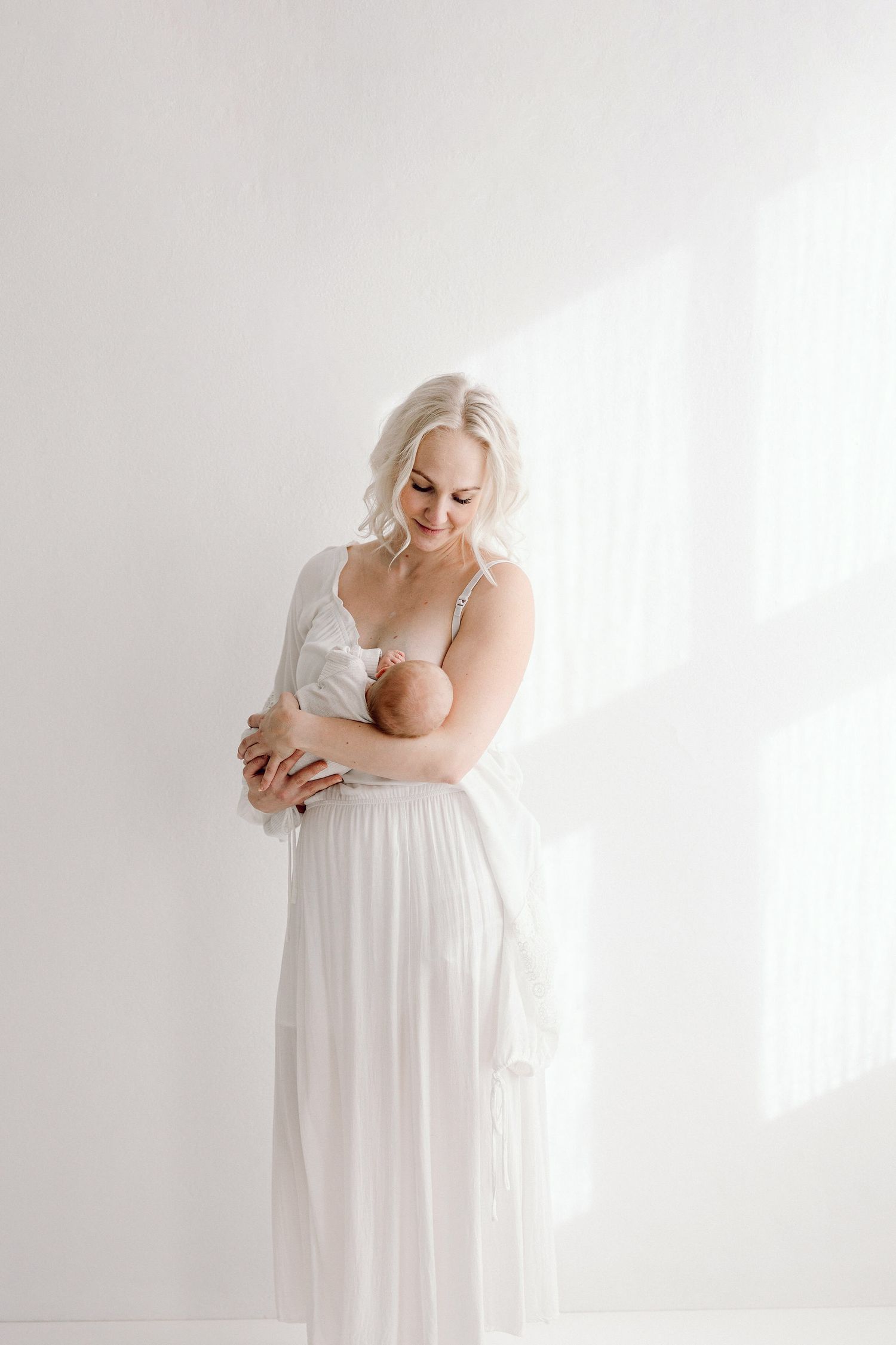 Mother in white dress holding baby in sunlit room.