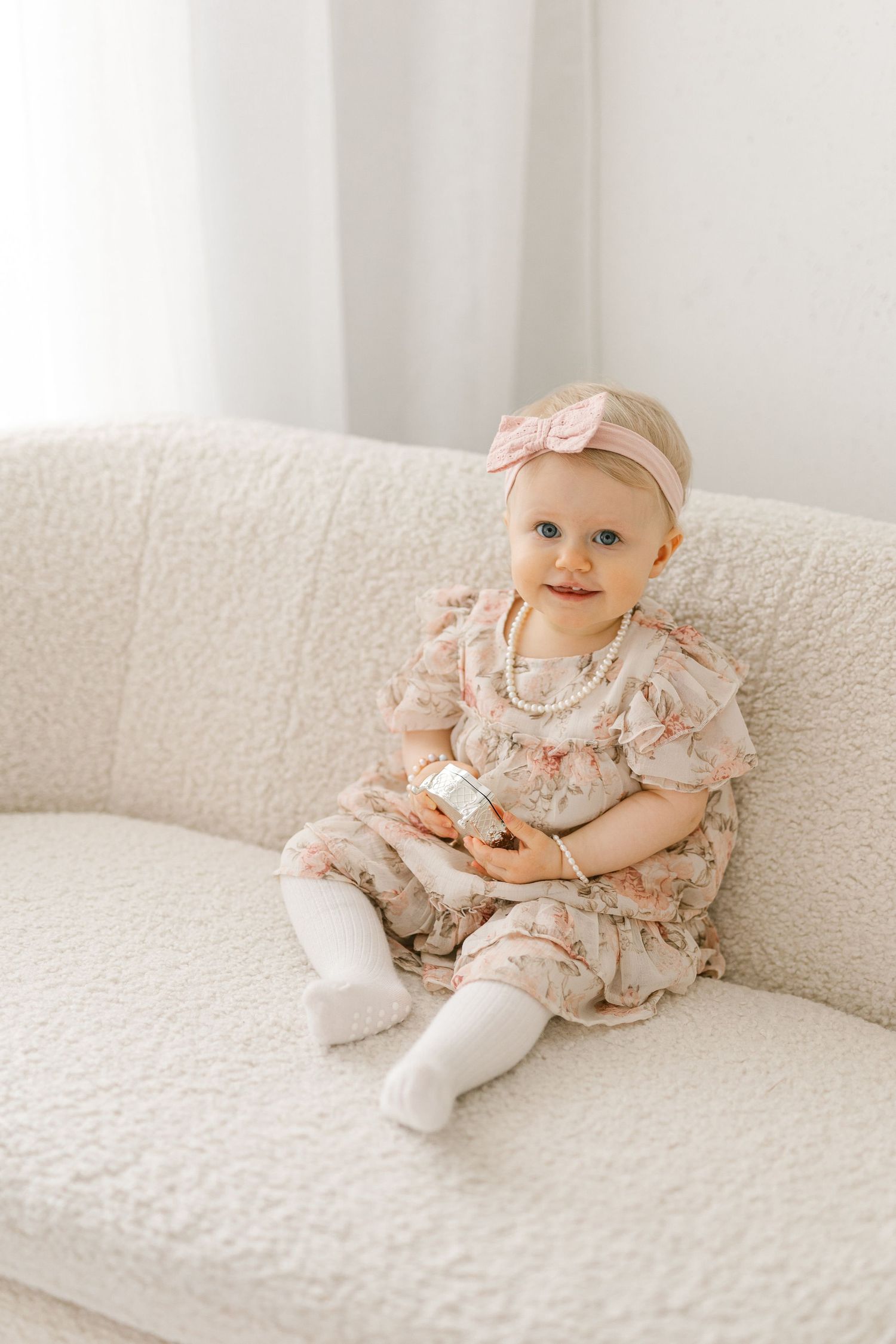 Baby wearing floral dress and bow headband sits smiling on white couch.