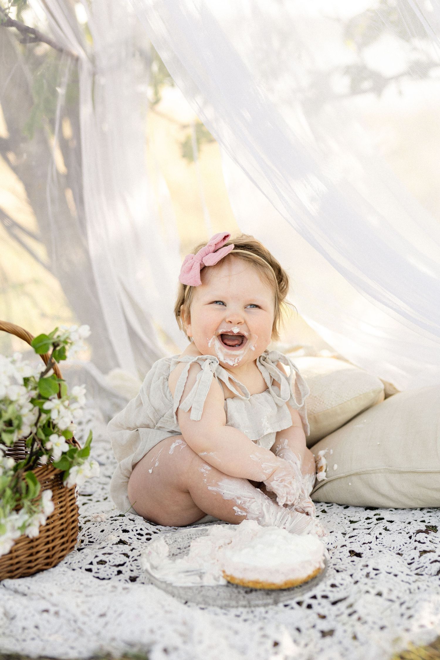 Baby in vintage dress sits on lace blanket next to picnic basket and flowers.