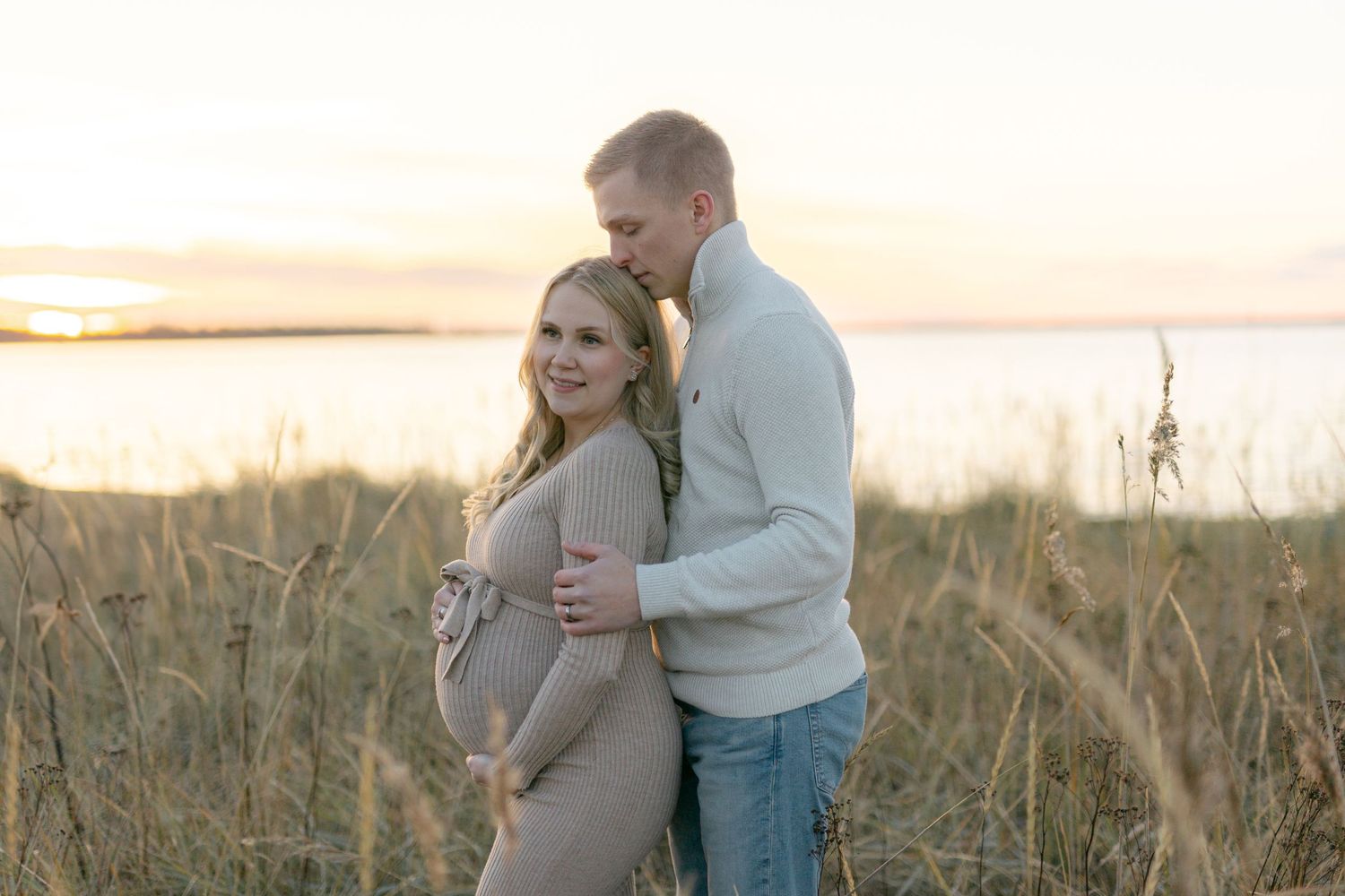Expectant couple embraces in a golden field at sunset, capturing a tender maternity moment.