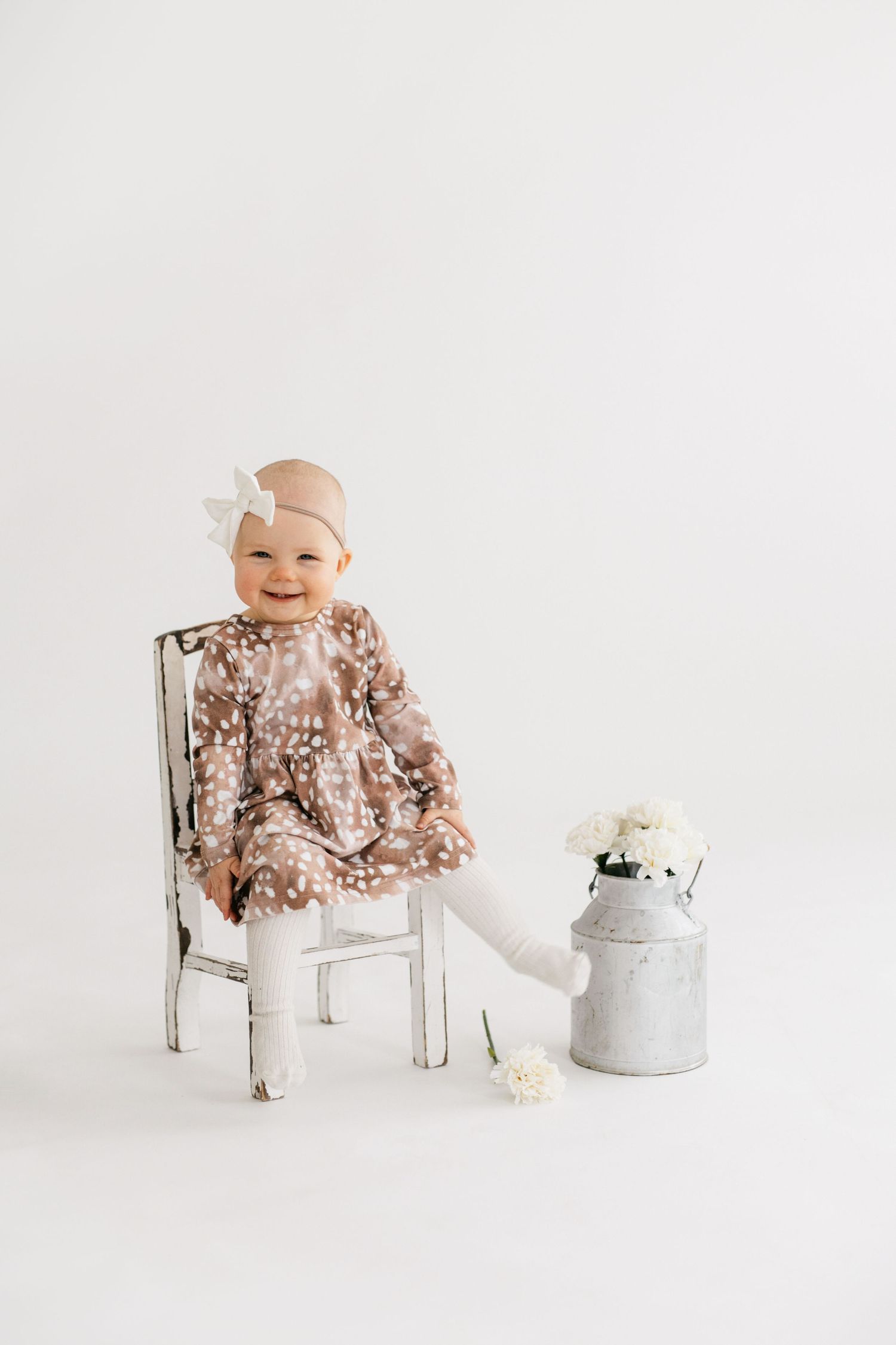 A happy child sits on a vintage white chair next to a metal milk can wearing a floral dress.