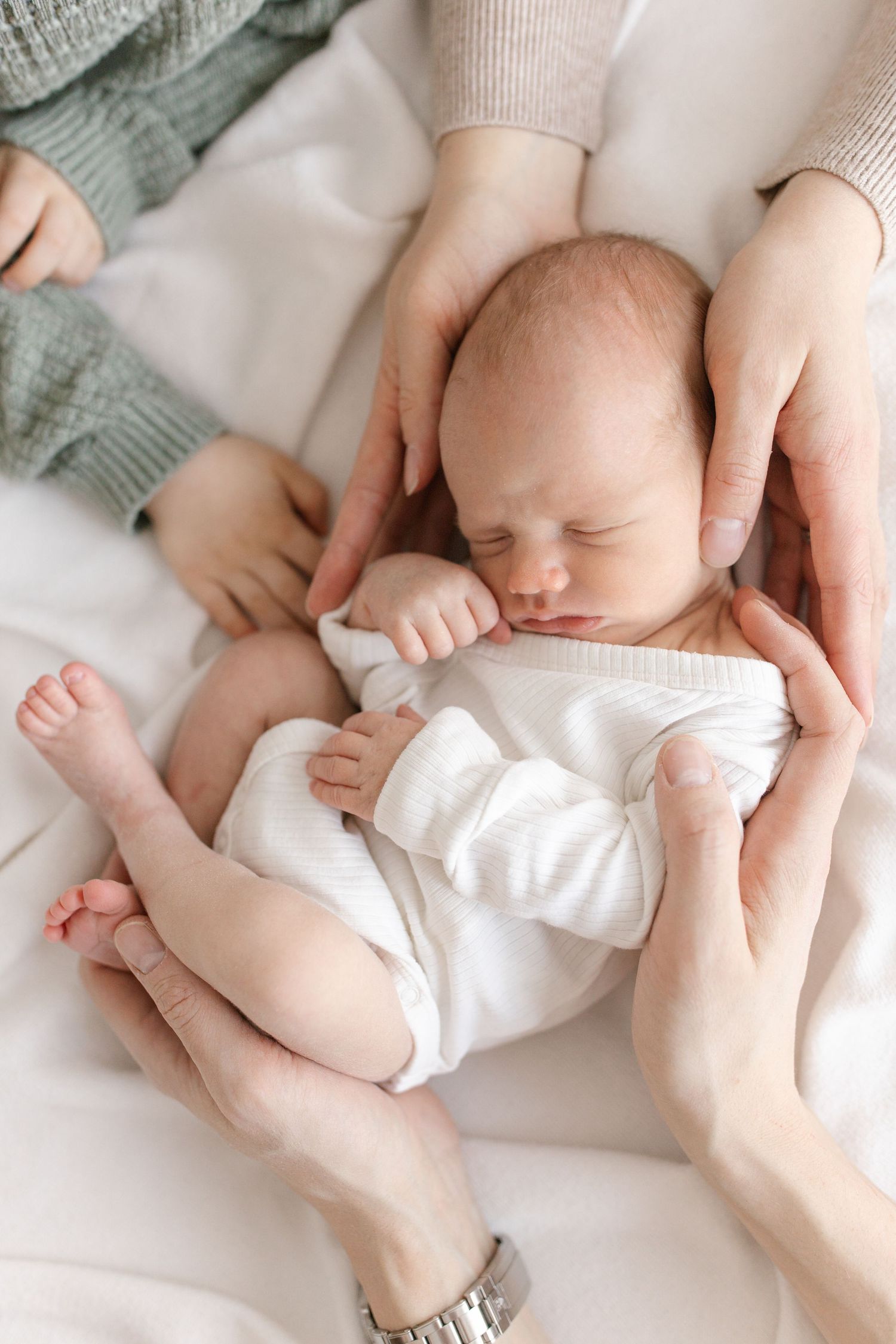 Newborn baby sleeping peacefully in parents hands on white blanket, wearing white onesie.