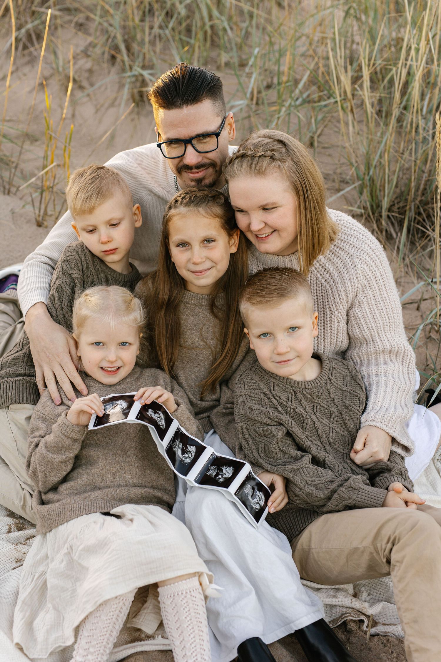 A family group wearing coordinated neutral sweaters and clothing sits together on a sandy beach near tall grass.