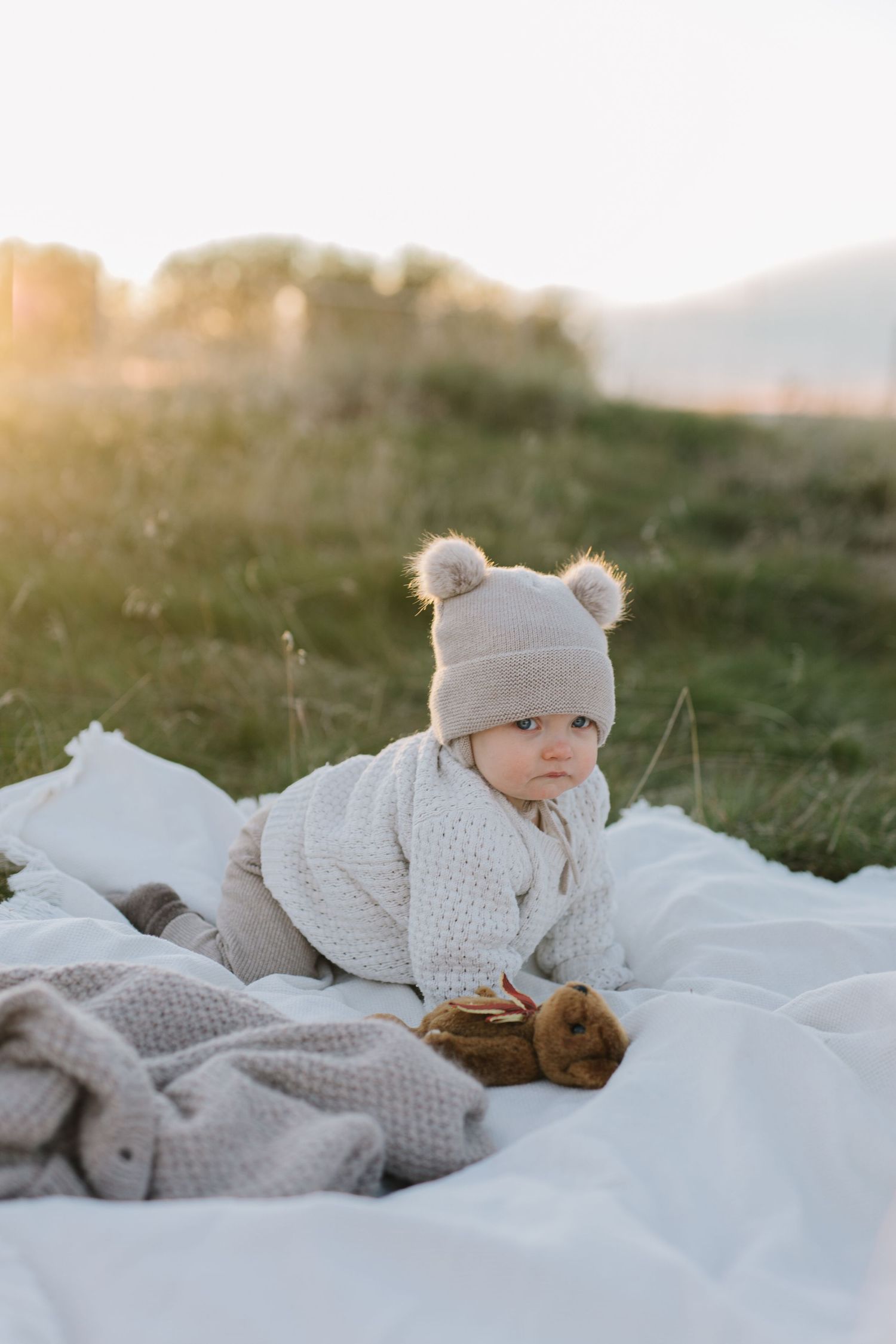 Baby in knit sweater and bear hat lying on blanket during golden hour outdoors.