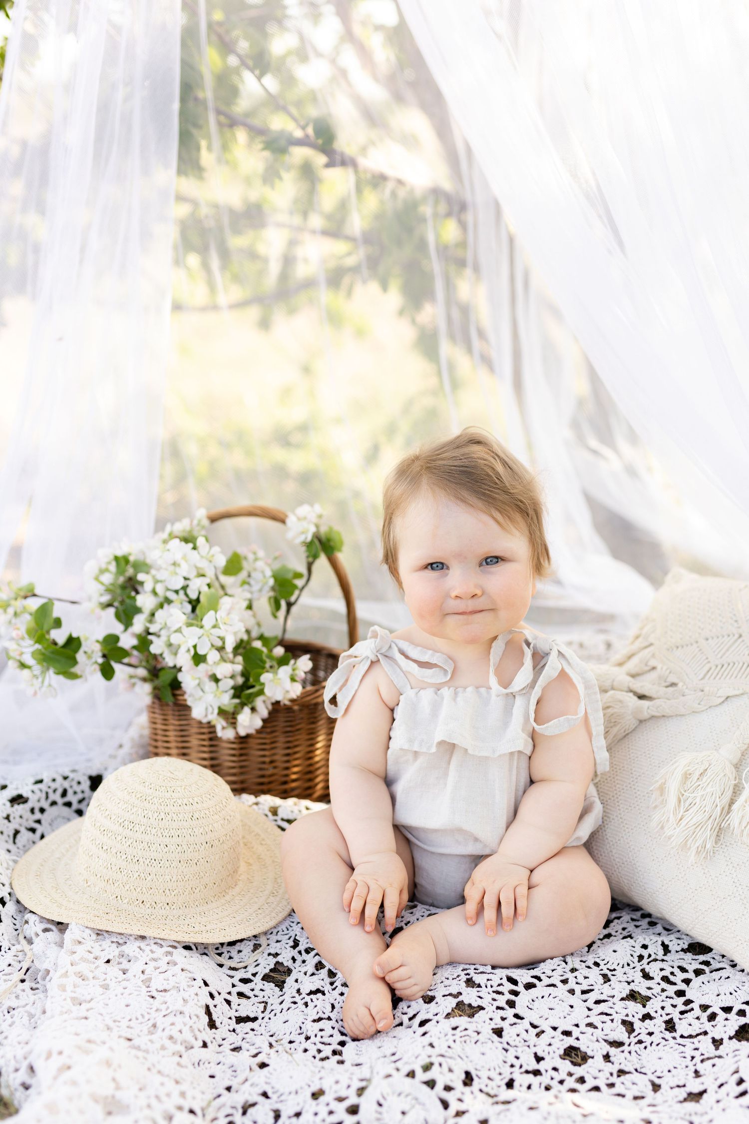 Baby in white outfit sits on lace blanket next to straw hat and flower basket outdoors.