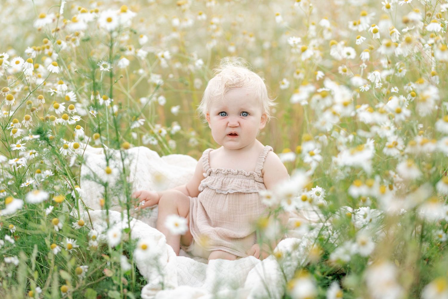 Baby in pink dress stands among blooming white daisies in a summer meadow.