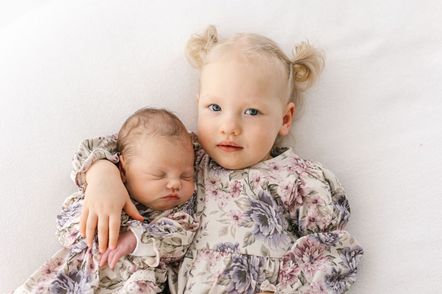 Two sleepy young children in floral dresses cuddle together on a soft white background.