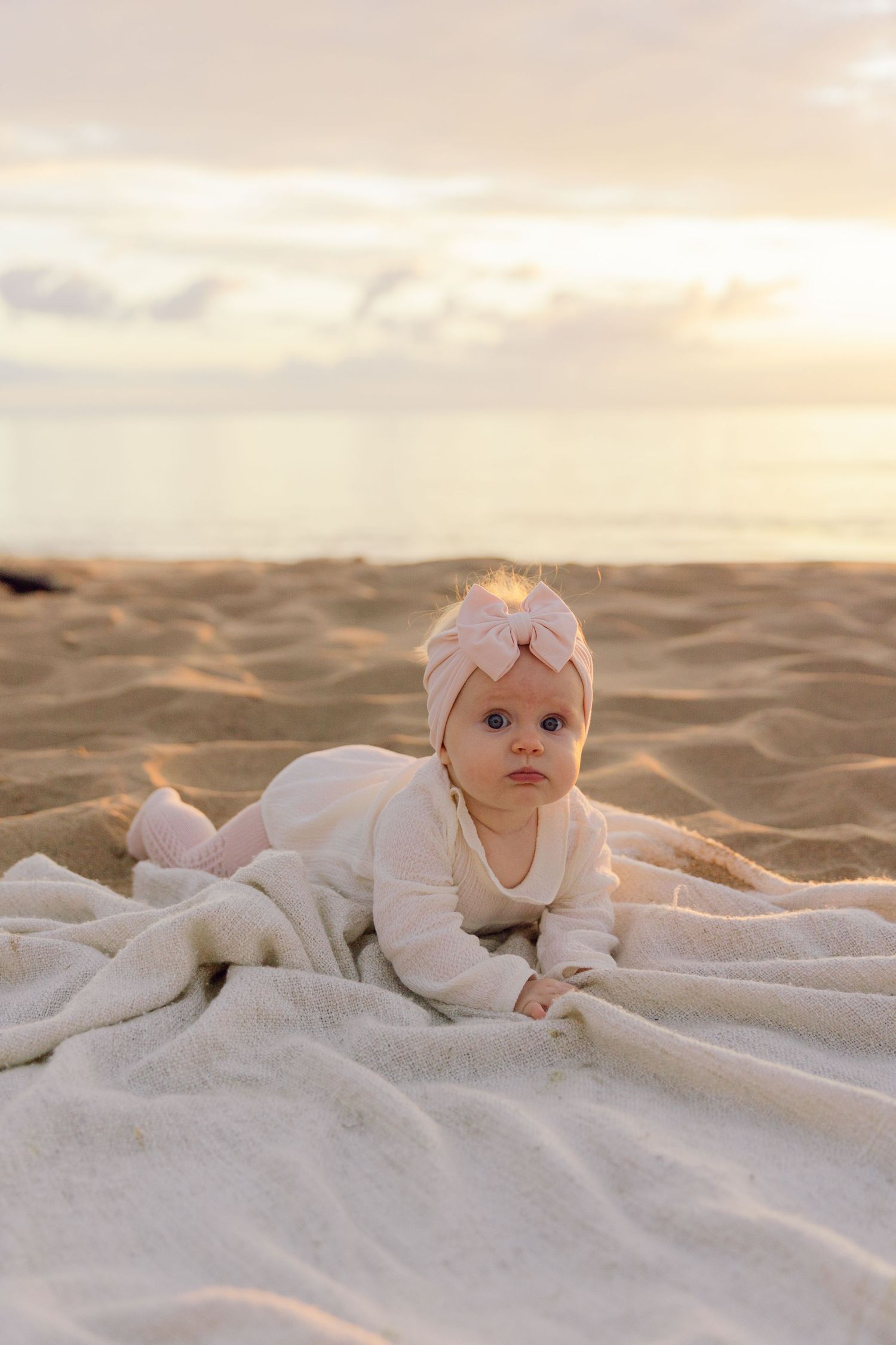 Baby in white outfit and headband lying on blanket at beach during sunset.