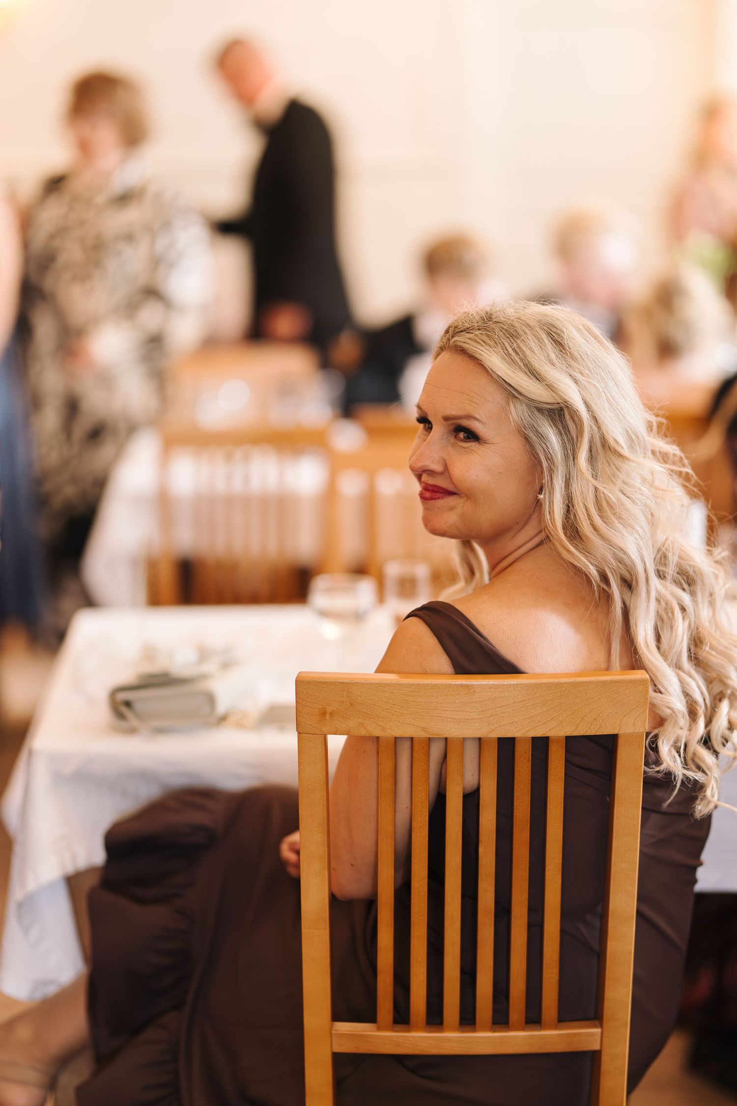 A woman with blonde wavy hair sits at a wooden chair during a social gathering.