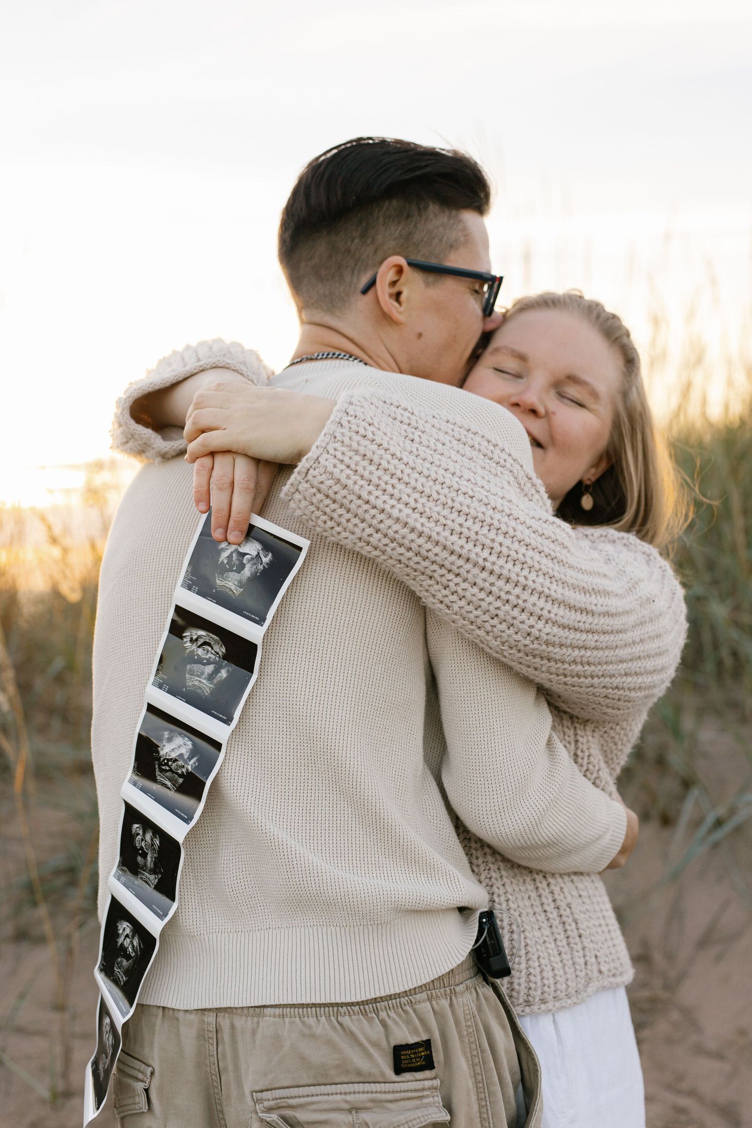 A couple embraces outdoors while holding ultrasound photos during a sunset pregnancy announcement photoshoot.