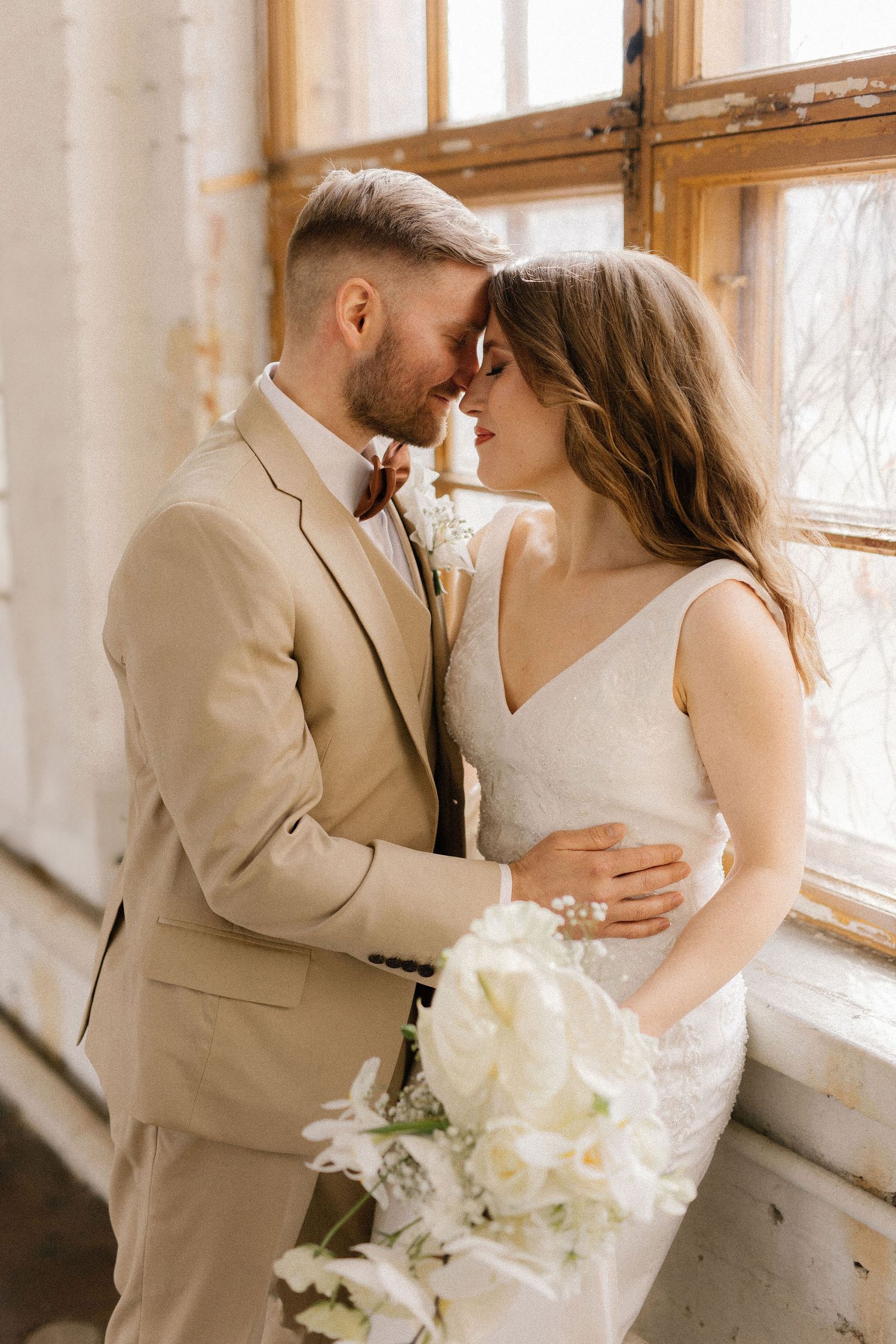 Wedding portrait showing tender moment between newlyweds with white floral bouquet near sunny windows.