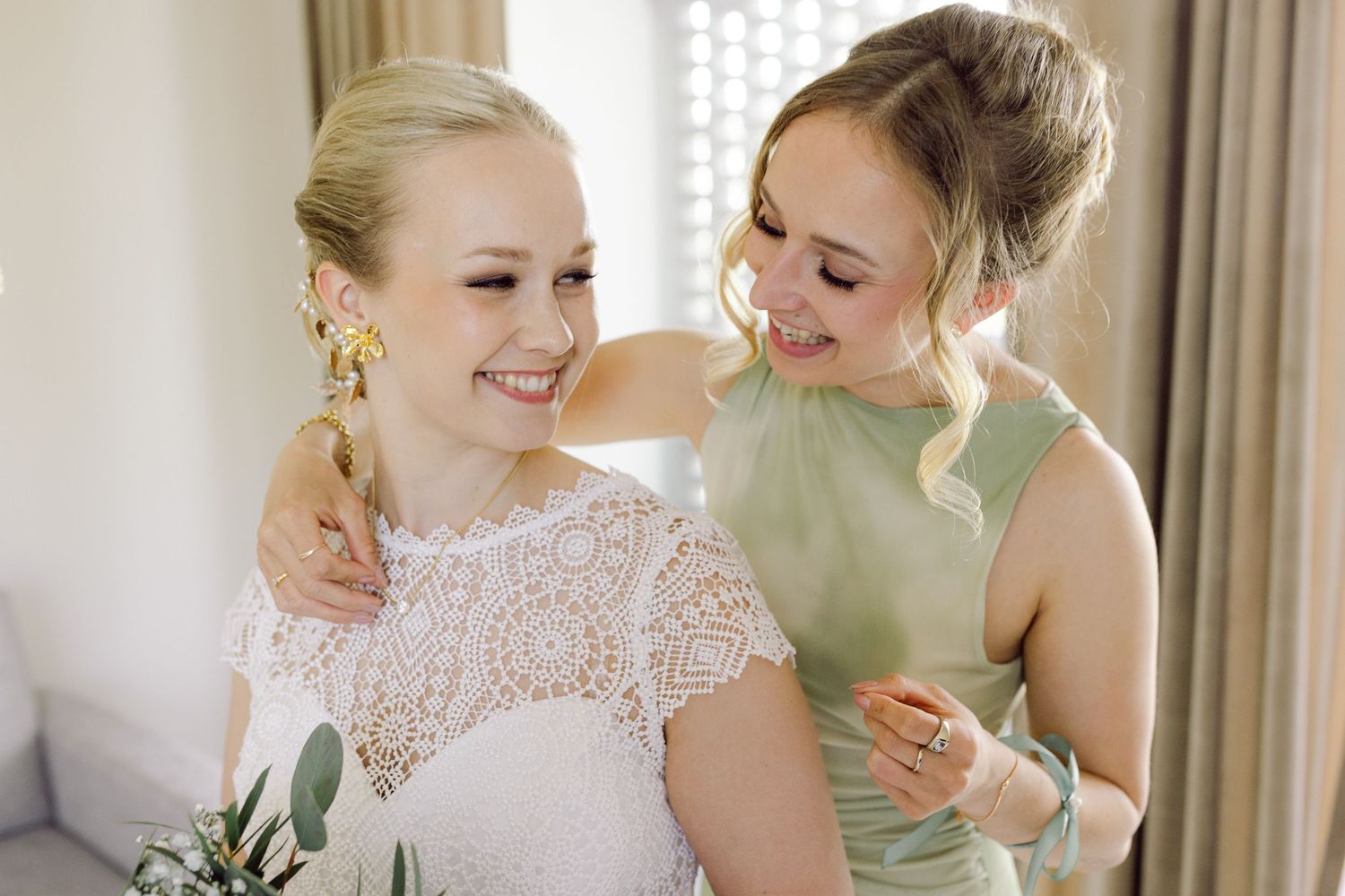 A bride and her bridesmaids share joyful moments while getting ready in a sunlit room.