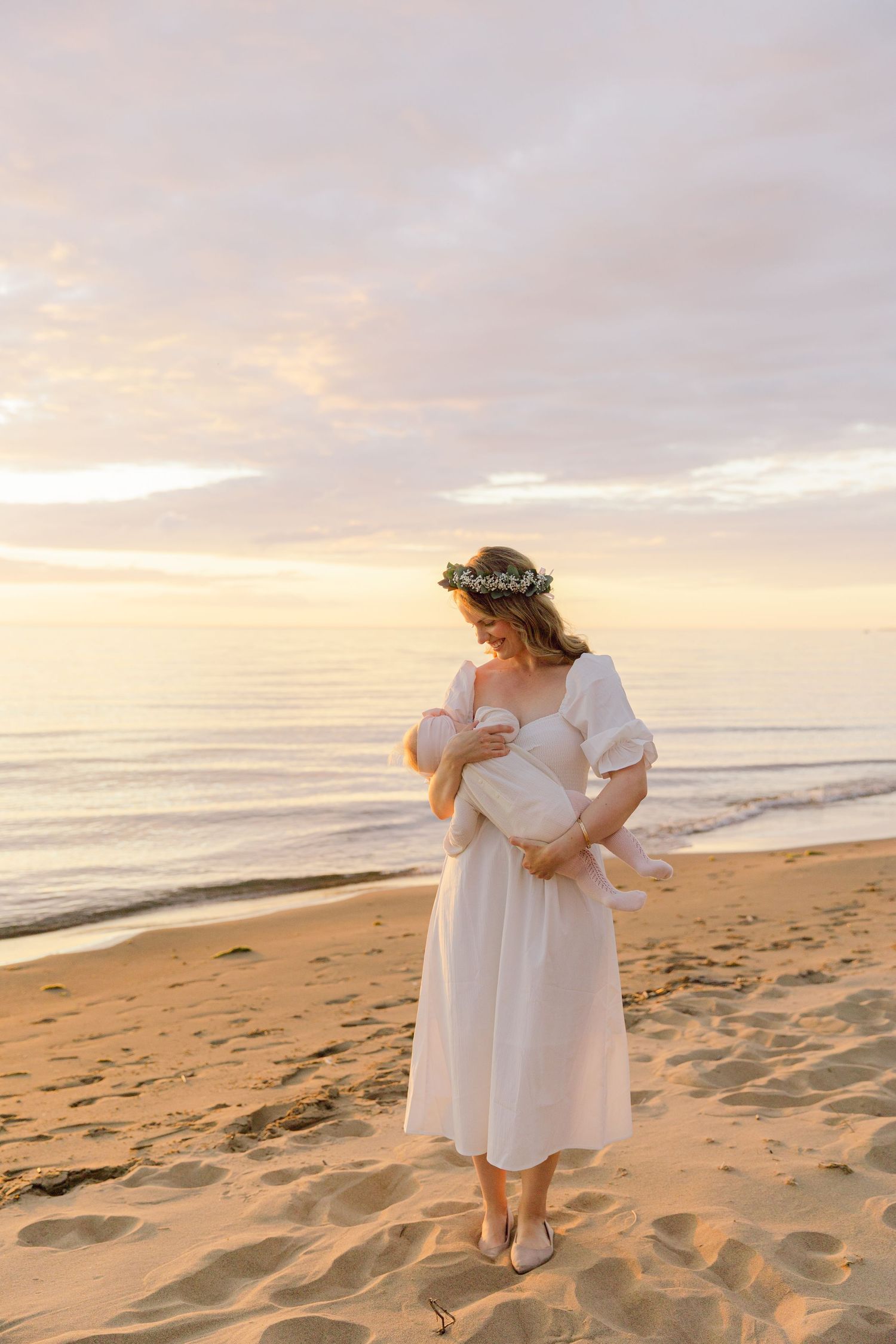 Woman in white dress holding baby on sandy beach during sunset.